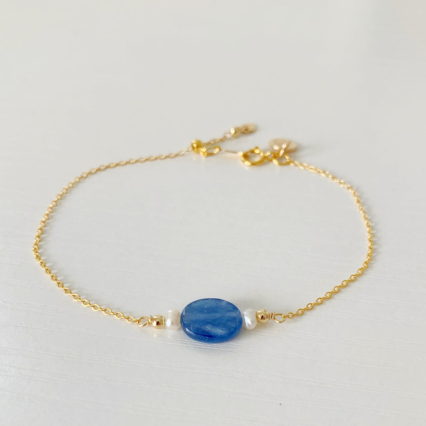 the brewster adjustable bracelet by mermaids and madeleines has a blue kyanite coin gem at the center with a petite freshwater pearl on either side, the bracelet has 14k gold filled chain, findings and adjustable slide bead clasp. this bracelet is photographed flat on a  white surface