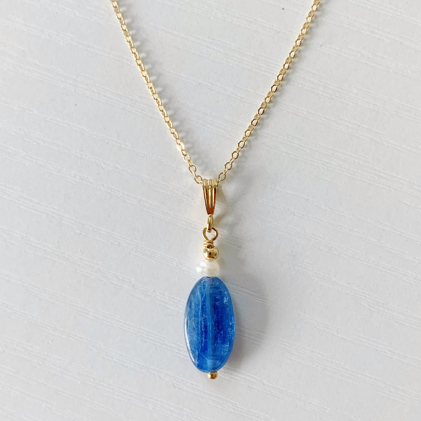 the brewster pendant by mermaids and madeleines is an elongated blue kyanite bead drop with a small freshwater pearl on top. the pendant is hanging from a 14k gold filled chain and findings. this necklace is photographed as a flatlay, closer up and laying on a white surface