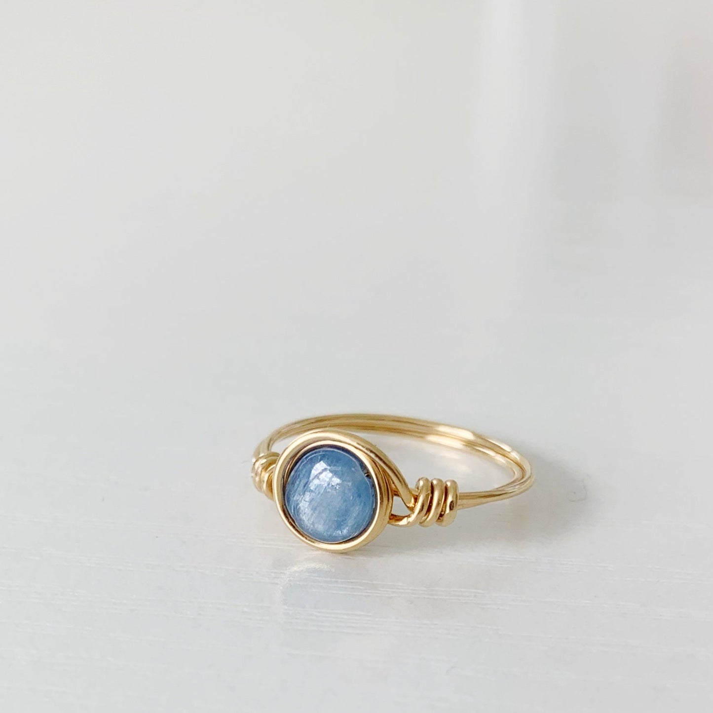 the brewster wire wrapped ring by mermaids and madeleines is a blue kyanite coin bead wrapped in 14k gold filled wire. this ring is photographed on a white surface