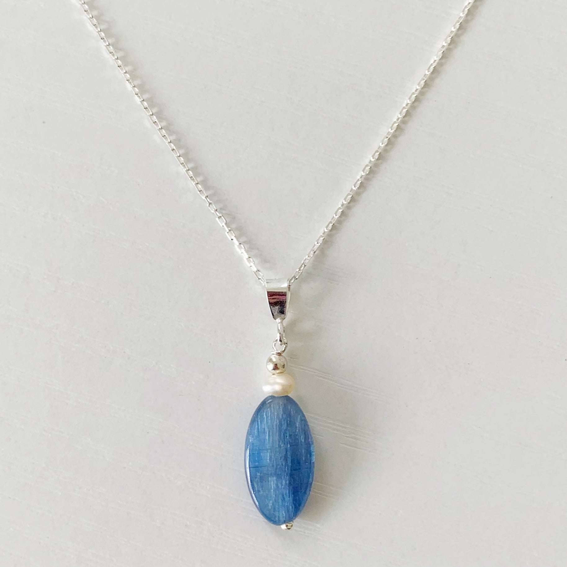 the brewster pendant by mermaids and madeleines is designed with an elongated blue kyanite oval and freshwater pearl drop suspended from a sterling silver chain. this necklace is photographed closer up as a flatlay on a white surface