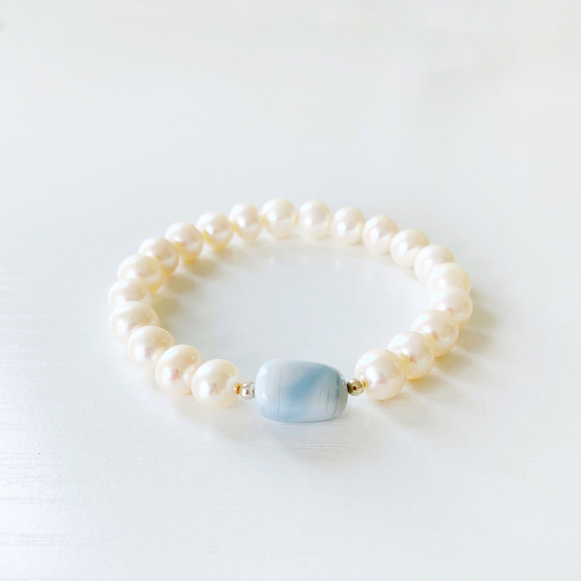 the blue hydrangea stretch bracelet by mermaids and madeleines is designed with a blue opal pebble bead at the center with freshwater pearls along the rest of the bracelet. this bracelet is photographed flat on a white surface