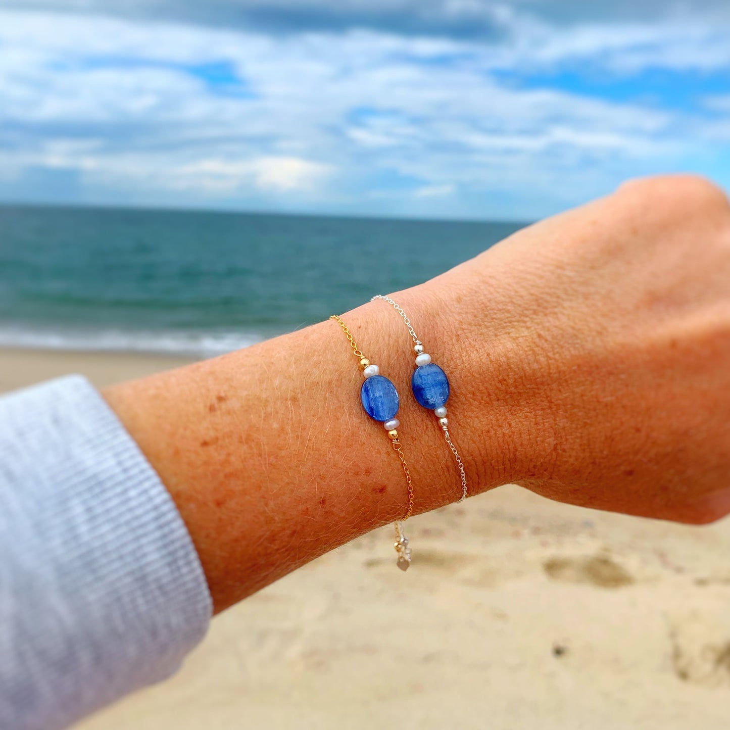 image shows 2 brewster bracelets stacked on a wrist. one bracelet in 14k gold filled and one in sterling silver. there is a soft beach scenery in the background