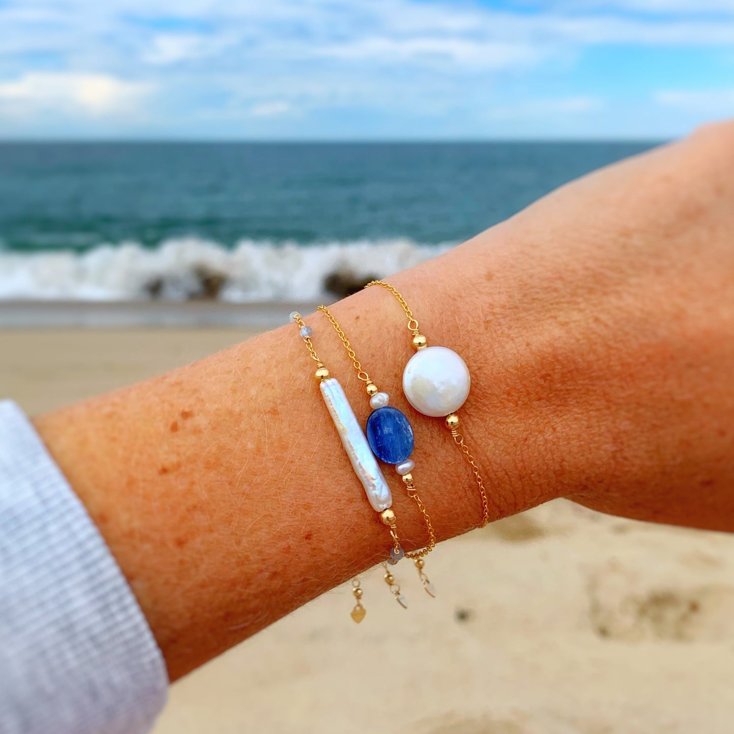 this is an image of a wrist with 3 of the mermaids and madeleines gold filled adjustable bracelets stacked on it with an ocean in the background. the bracelets from left to right are the truro, the brewster and the newport bracelet