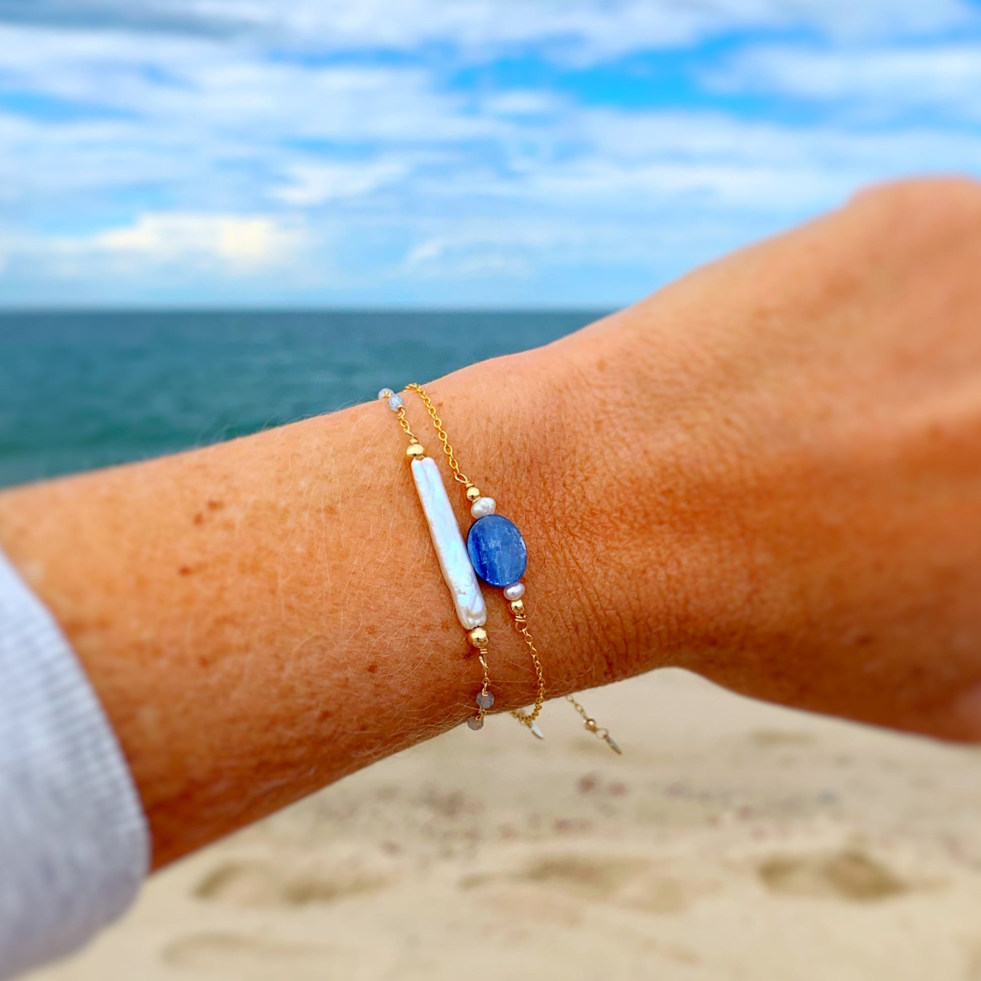 this image shows 2 bracelets close up stacked on a wrist. the first bracelet is the truro bracelet and the second is the brewster bracelet both in 14k gold filled. there is a soft beach scenery background