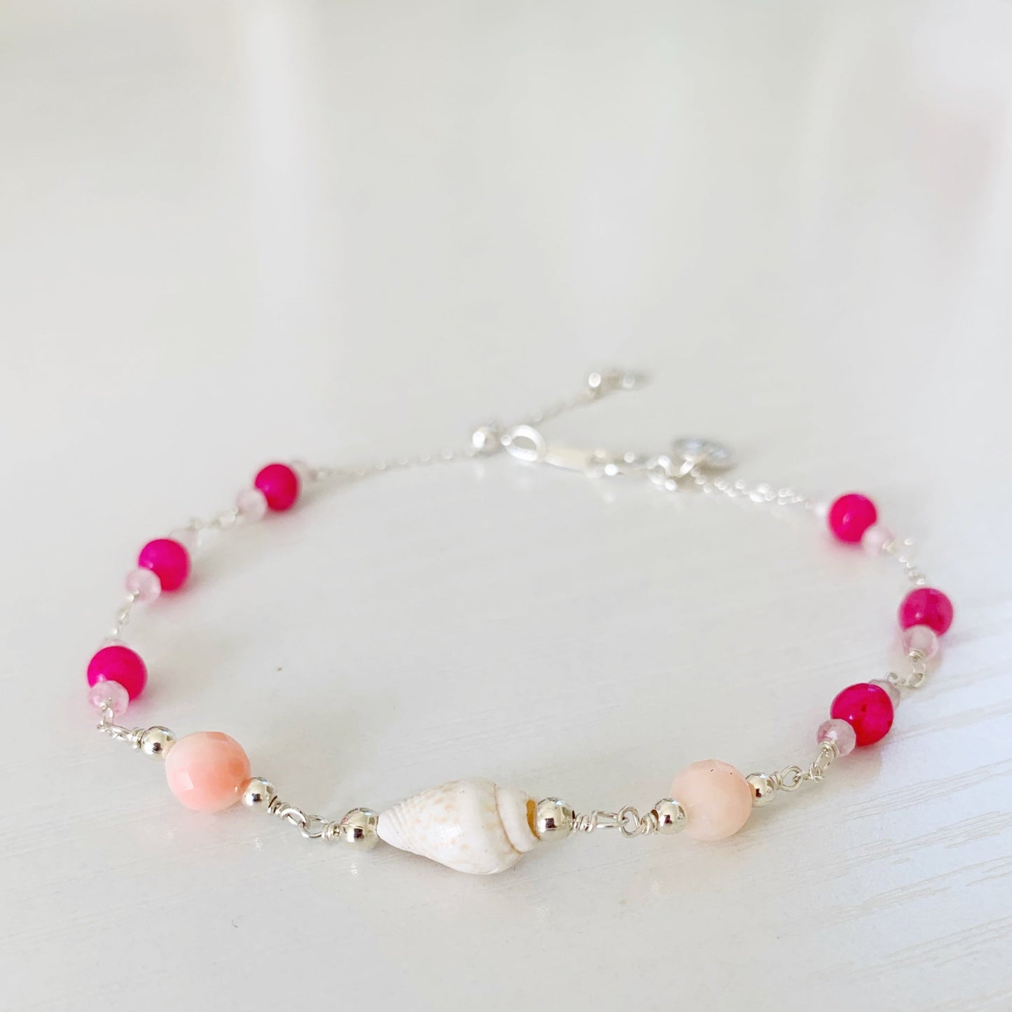 the pink flamingo shellebration bracelet by mermaids and madeleines is a sterling silver and beaded adjustable bracelet with a shell and coral beads at the center complimented by alternating dyed pink jade, rose quartz. this bracelet is photographed on a white surface