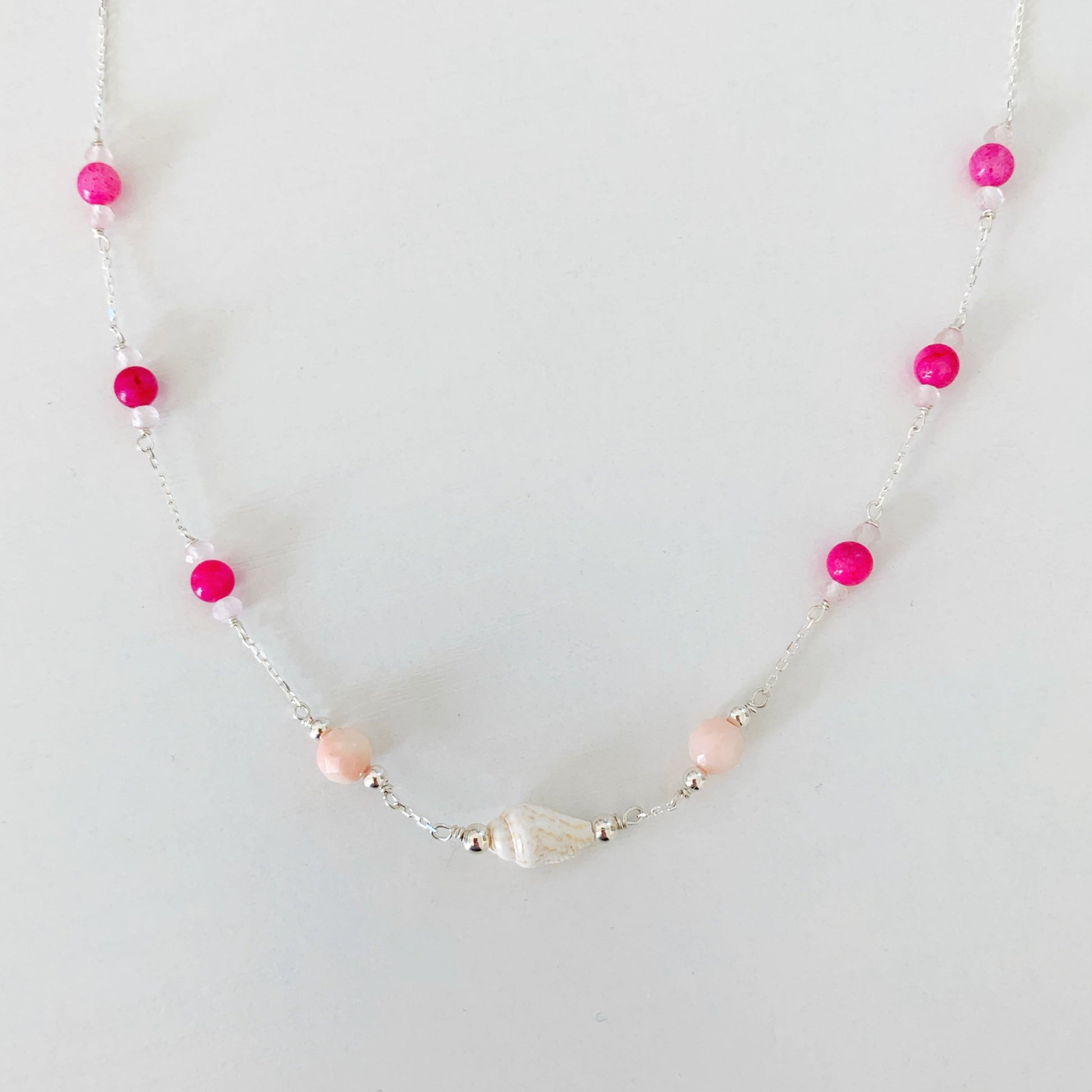 The pink flamingo shellebration necklace by mermaids and madeleines is created with a shell and coral beads at the center with dyed hot pink jade and rose quartz rondelles spaced out going up the sterling chain to the clasp. this necklace is photographed on a white surface