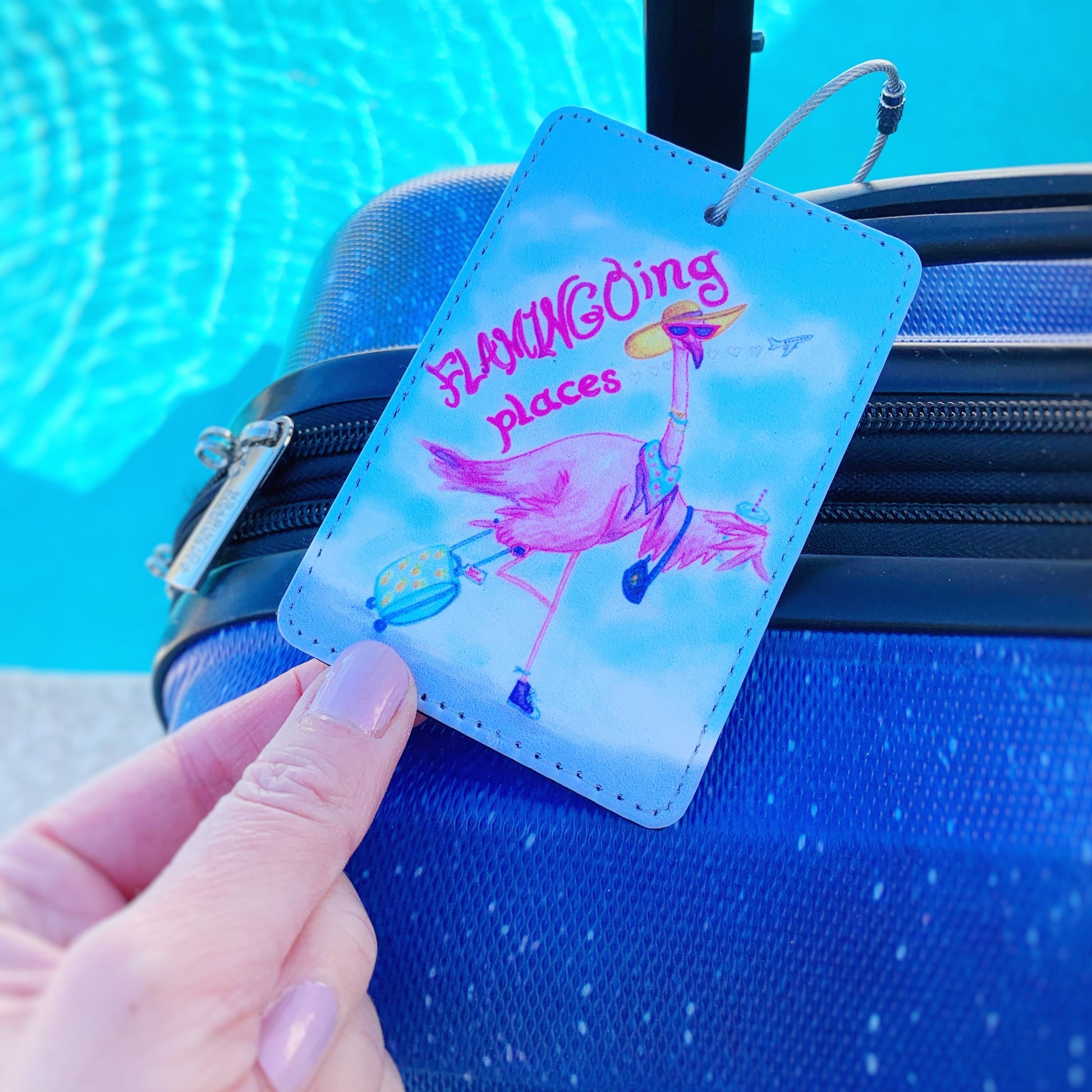 flamingal luggage tag by mermaids and madeleines. this tag has a bright pink flamingo on it carrying a suitcase. this tag is photographed close up on a suitcase with a hand tilting it toward the camera.