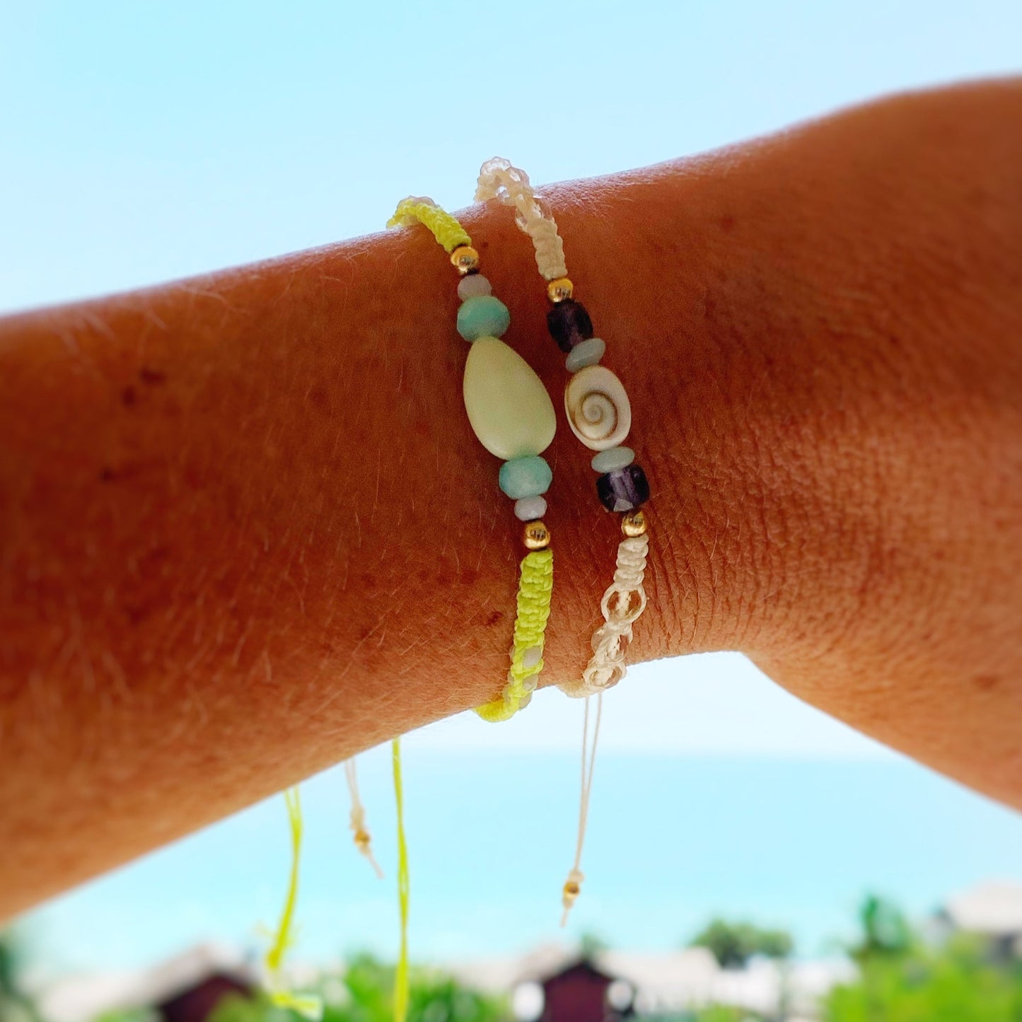 this image is a photo of a wrist with 2 macrame bracelets stacked and a light blue blurred beachy background. the 2 bracelets are macrame, the top one is white with a shiva shell at the center and is the mermaid money bracelet, the second bracelet is sips on the court, a neon yellow macrame bracelet