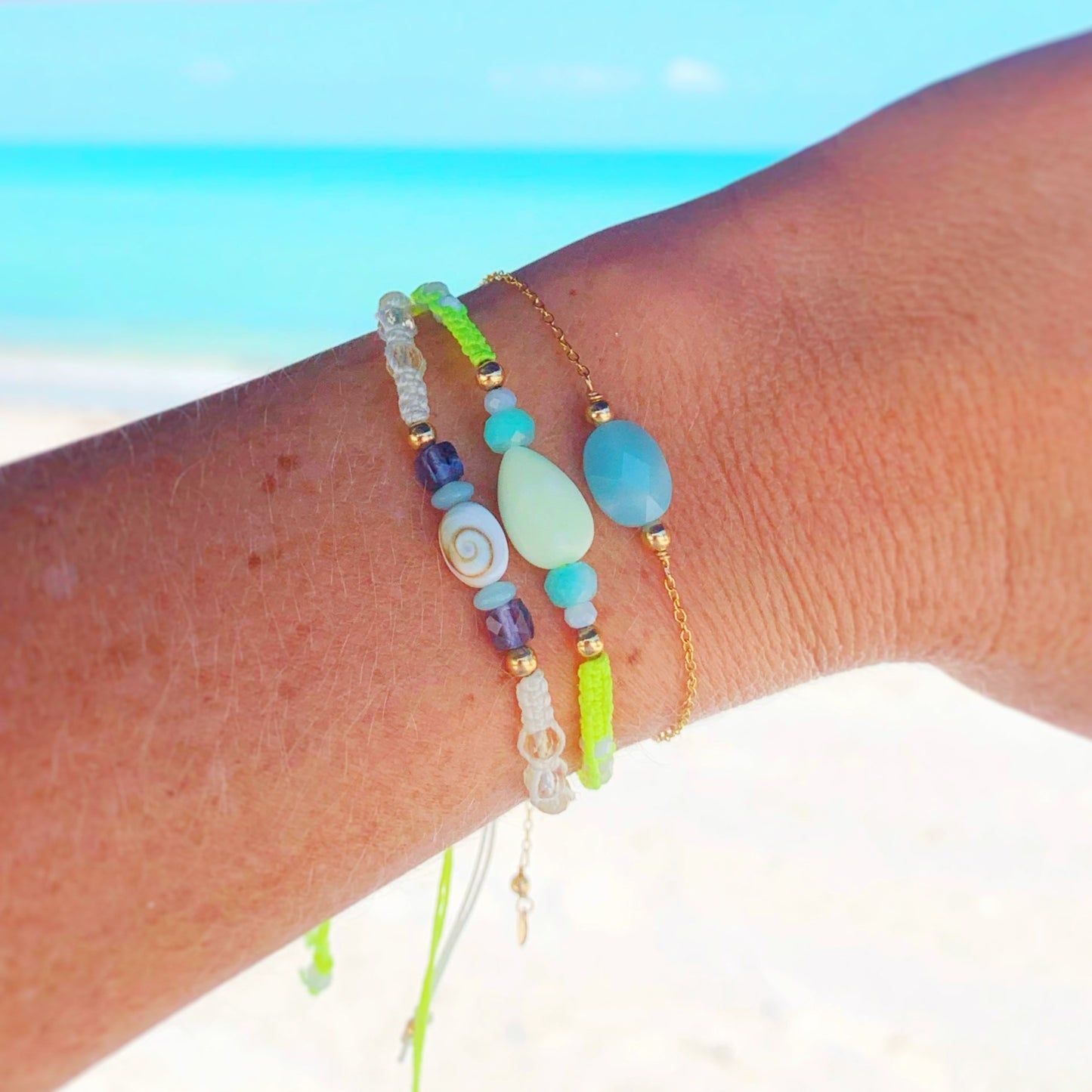 image of 3 mermaids and madeleines bracelet stacked up on a wrist with a washed out beach image in the background. the 3 bracelets on the wrist from left to right are the mermaid money macrame bracelet, the sips on the court neon yellow macrame bracelet and the laguna bracelet in gold filled