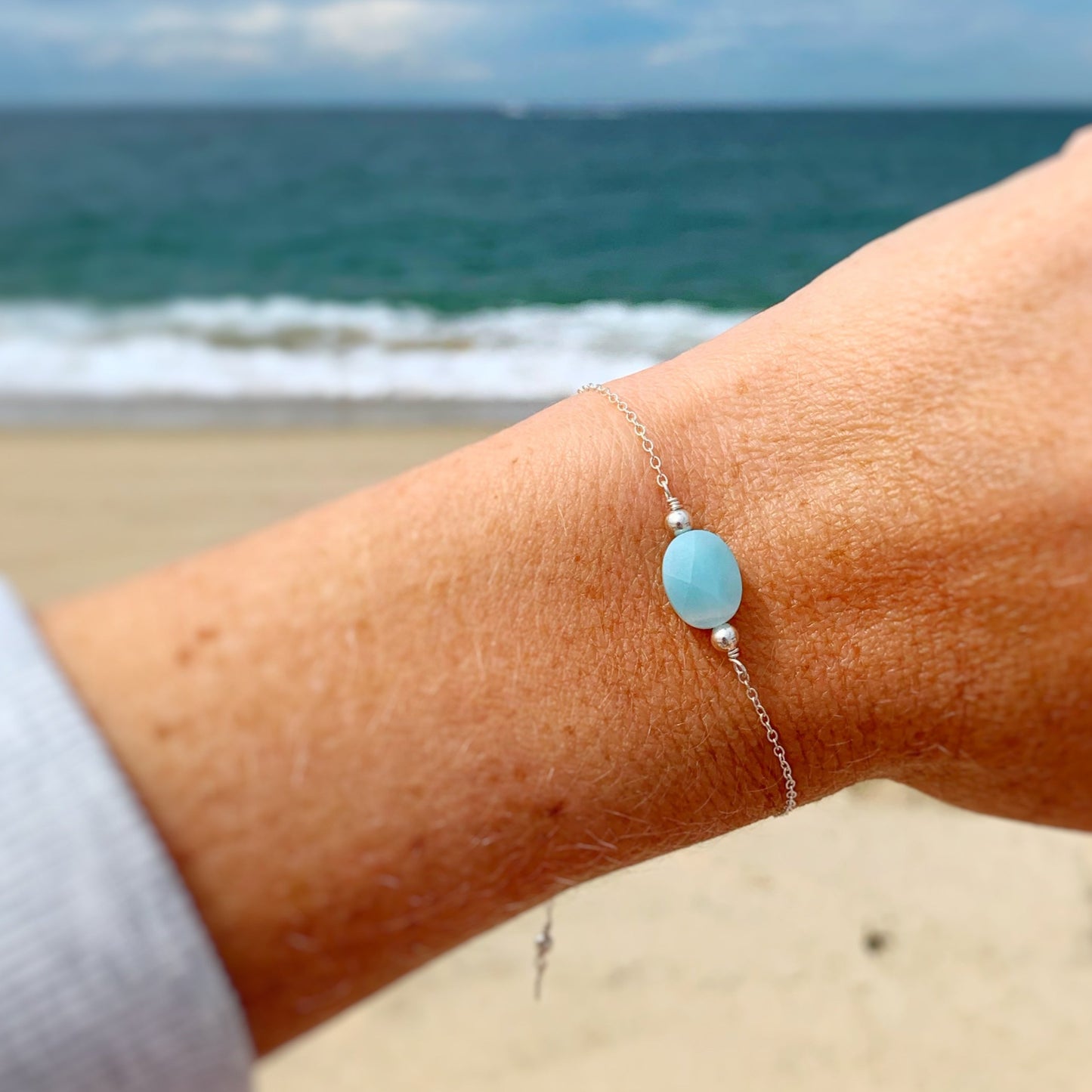 image of the mermaids and madeleines laguna bracelet on a wrist. this bracelet has an amazonite bead at the center with sterling silver chain. this bracelet is photographed on a wrist with a beach image in the background