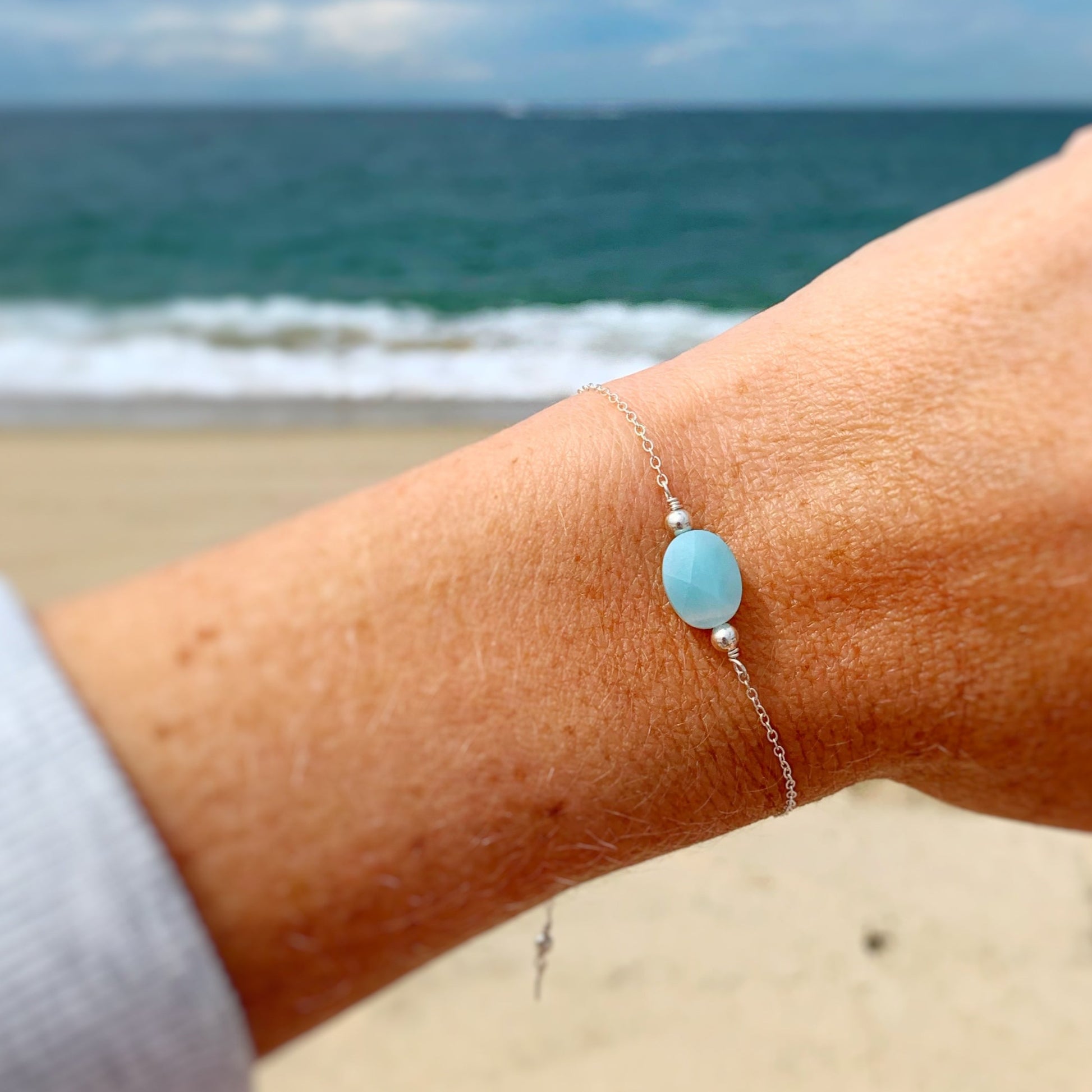 image of the mermaids and madeleines laguna bracelet on a wrist. this bracelet has an amazonite bead at the center with sterling silver chain. this bracelet is photographed on a wrist with a beach image in the background