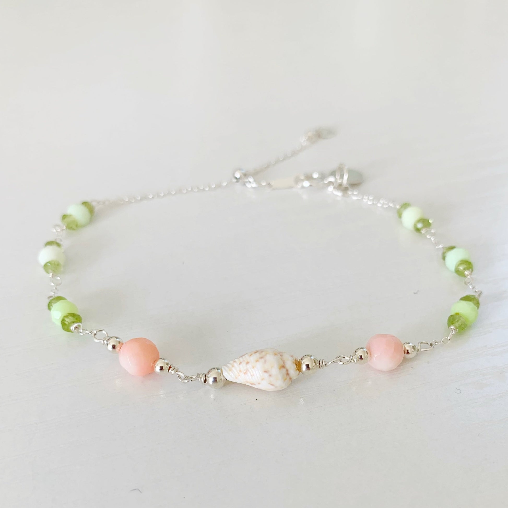 the margarita shellebration bracelet by mermaids and madeleines is a sterling silver and beaded adjustable bracelet with a shell and coral beads at the center complimented by alternating green opal and peridot. this bracelet is photographed on a white surface