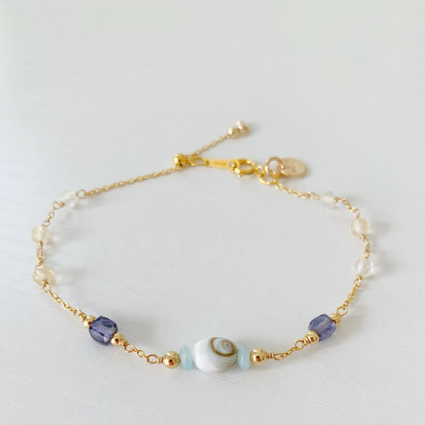 the mermaid money adjustable bracelet is a 14k gold filled chain bracelet with an adjustable slide bead clasp by the spring ring. the center of the bracelet has a shiva shell and on either side is an amazonite saucer bead . along the sides are an iolite cube, and round faceted citrine beads. this bracelet is photographed flat on a white surface