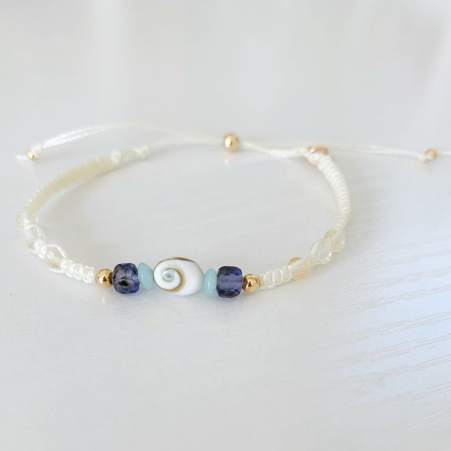 the mermaid money macrame bracelet by mermaids and madeleines is a cream color macrame bracelet with a shiva shell at the center and amazonite, iolite and 14k gold beads on either side and three faceted citrine beads on each side. This bracelet has a 14k gold filled slide bead as a clasp. The bracelet is photographed on a white surface