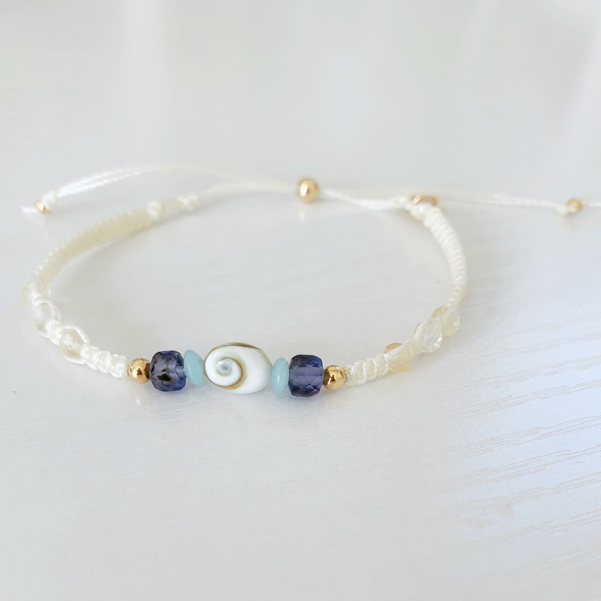 the mermaid money macrame bracelet by mermaids and madeleines is a cream color macrame bracelet with a shiva shell at the center and amazonite, iolite and 14k gold beads on either side and three faceted citrine beads on each side. This bracelet has a 14k gold filled slide bead as a clasp. The bracelet is photographed on a white surface