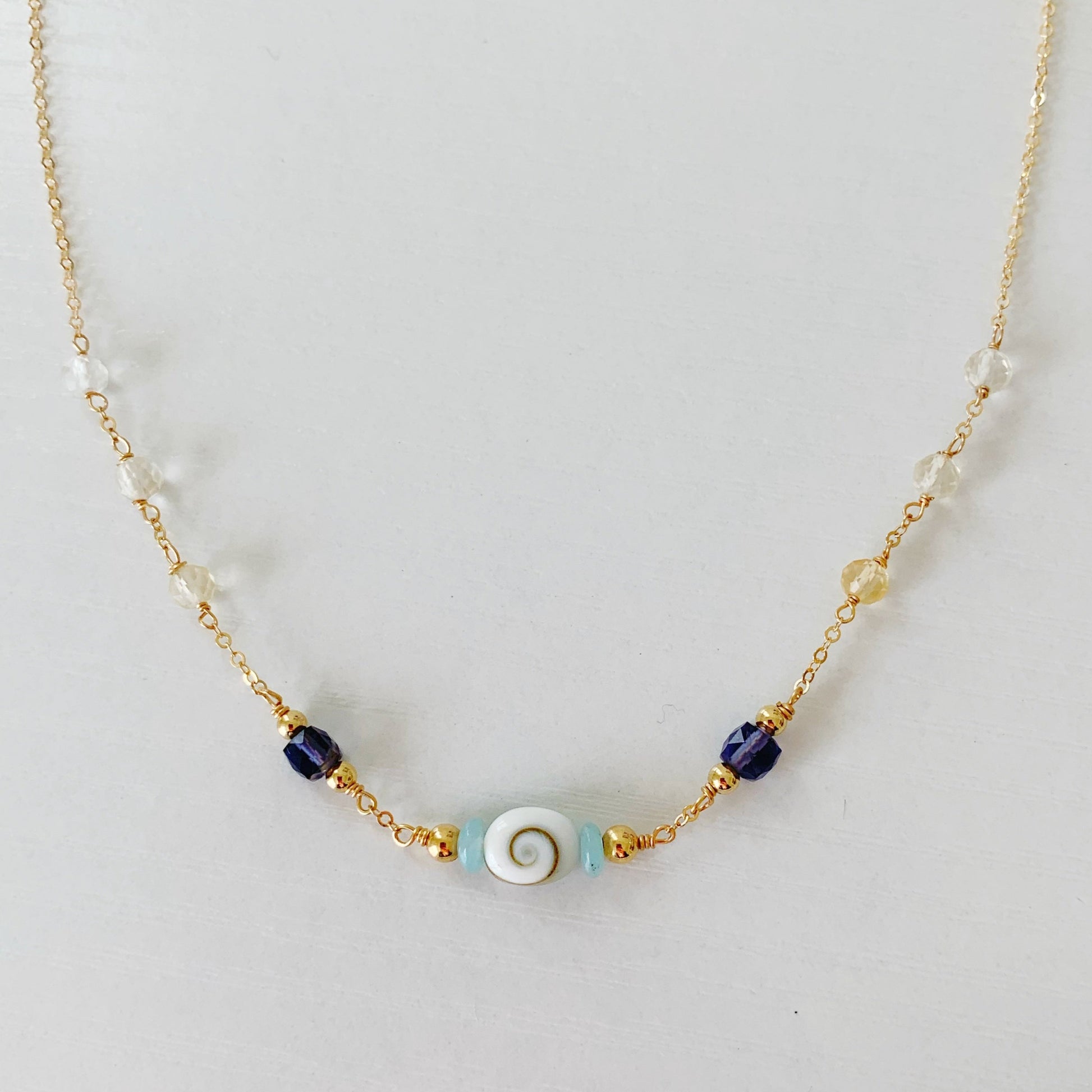 Mermaid Money Necklace by mermaids and madeleines is created with a chain and bead style necklace, a shiva shell oval bead at center complimented by amazonite disc and iolite faceted cube beads on either side. all combined with 14k gold filled beads, chain and findings. this necklace is photographed on a white background