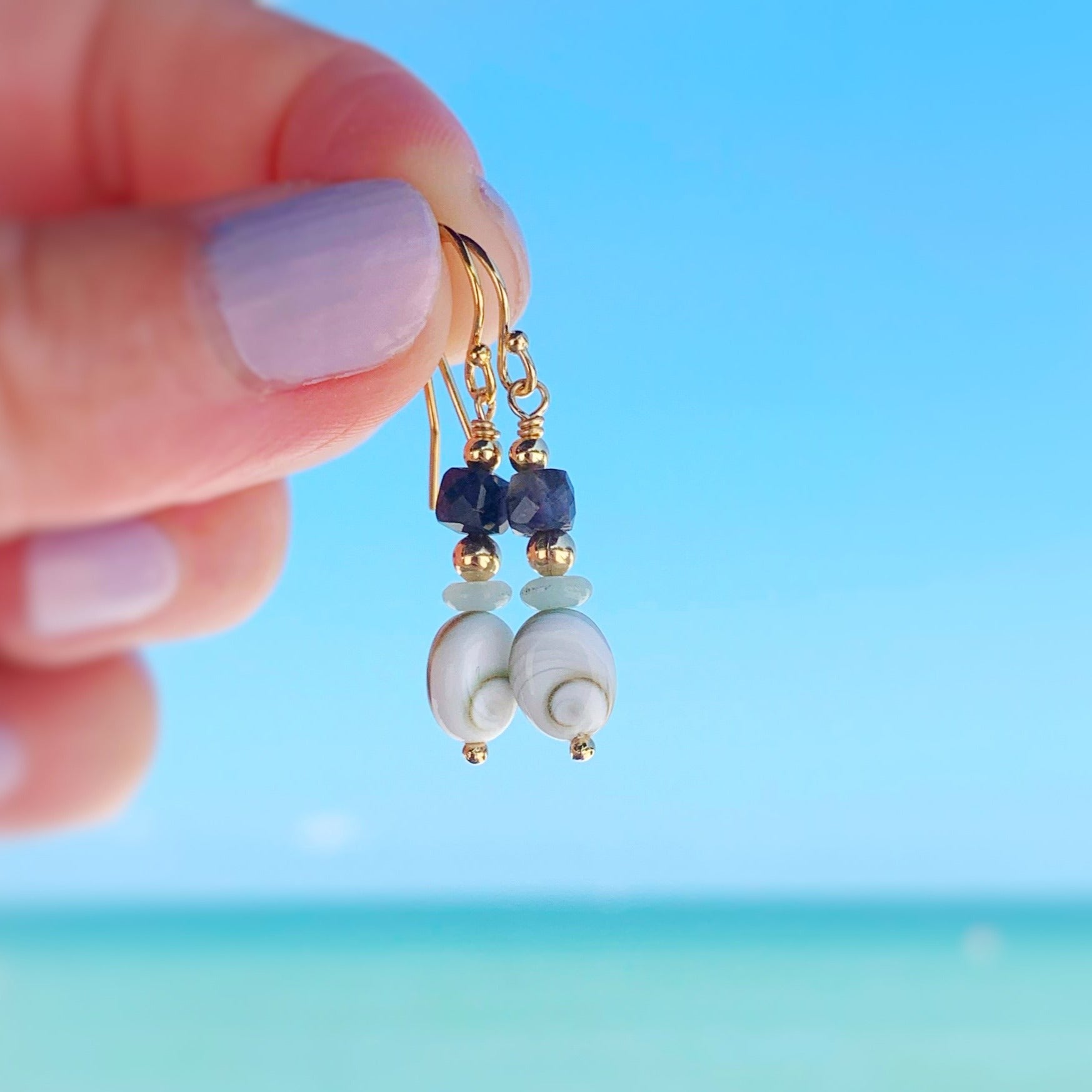 Mermaid Money earrings by mermaids and madeleines are designed with shiva shell ovals, small amazonite disc beads and faceted iolite cubes with 14k gold filled findings. this pair is held up in a hand with a blurred aqua blue beach scene in the background