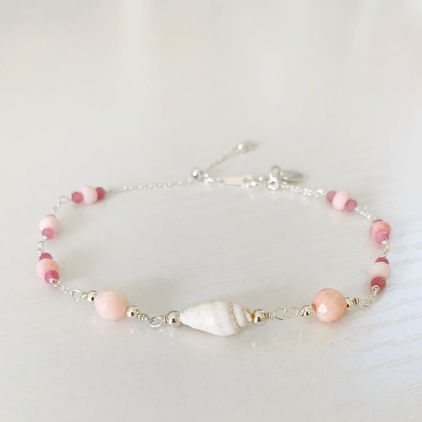 the pink lemonade shellebration bracelet by mermaids and madeleines is a sterling silver and beaded adjustable bracelet with a shell and coral beads at the center complimented by alternating pink opal and tourmaline. this bracelet is photographed on a white surface