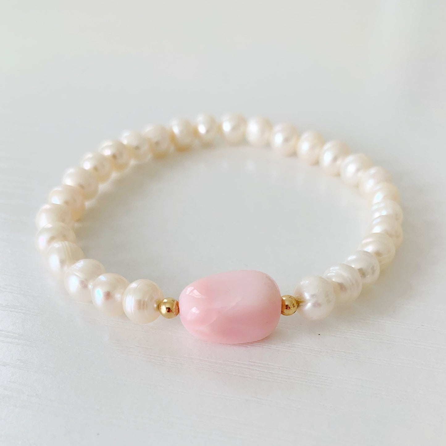 the pink peony stretch bracelet by mermaids and madeleines is a stretchy that features a pink opal nugget at the center with 14k gold filled 3mm round beads on either side and the rest of the bracelet is 7mm freshwater pearls. this bracelet is photographed flat on a whtie surface