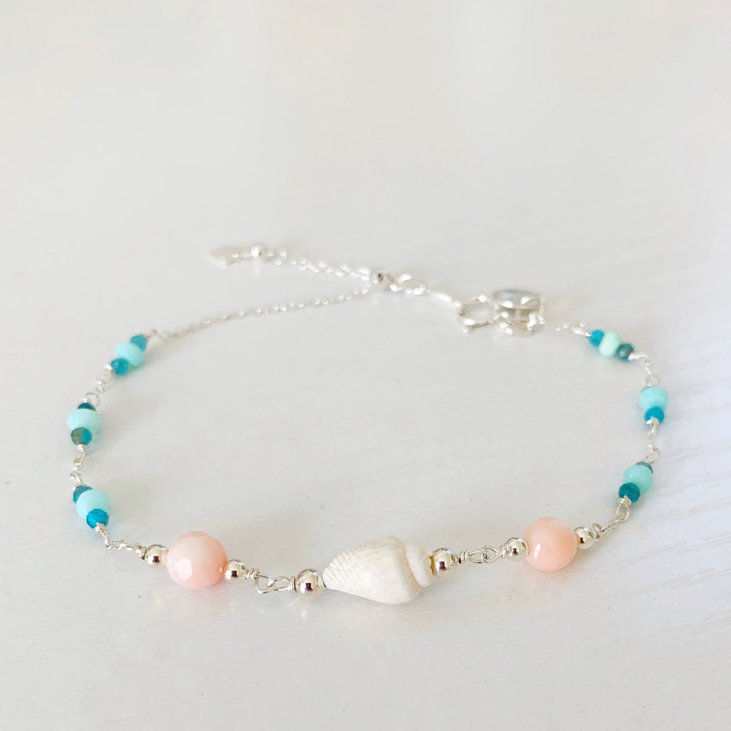 the pool party shellebration bracelet by mermaids and madeleines is a sterling silver and beaded adjustable bracelet with a shell and coral beads at the center complimented by alternating Peruvian opal and apatite. this bracelet is photographed on a white surface