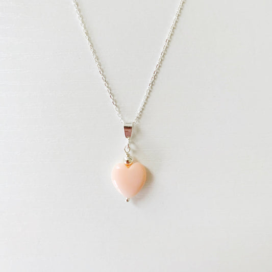 the pink posy necklace by mermaids and madeleines is a pendant style necklace on a sterling silver cable chain with length options of 16" or 18" and the pendant is a pink heart shaped conch shell. this necklace is photographed flat on a white surface with the front half of the necklace in view