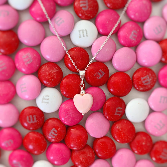 The pink posy necklace by mermaids and madeleines is a pendant style necklace with pink conch shell heart bead with sterling silver findings and chain. this  pendant is pictured over valentine's colored M+M candies
