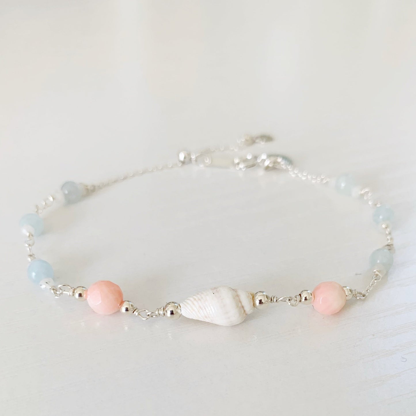 the seersucker shellebration bracelet by mermaids and madeleines is a sterling silver and beaded adjustable bracelet with a shell and coral beads at the center complimented by alternating aquamarine and moonstone. this bracelet is photographed on a white surface