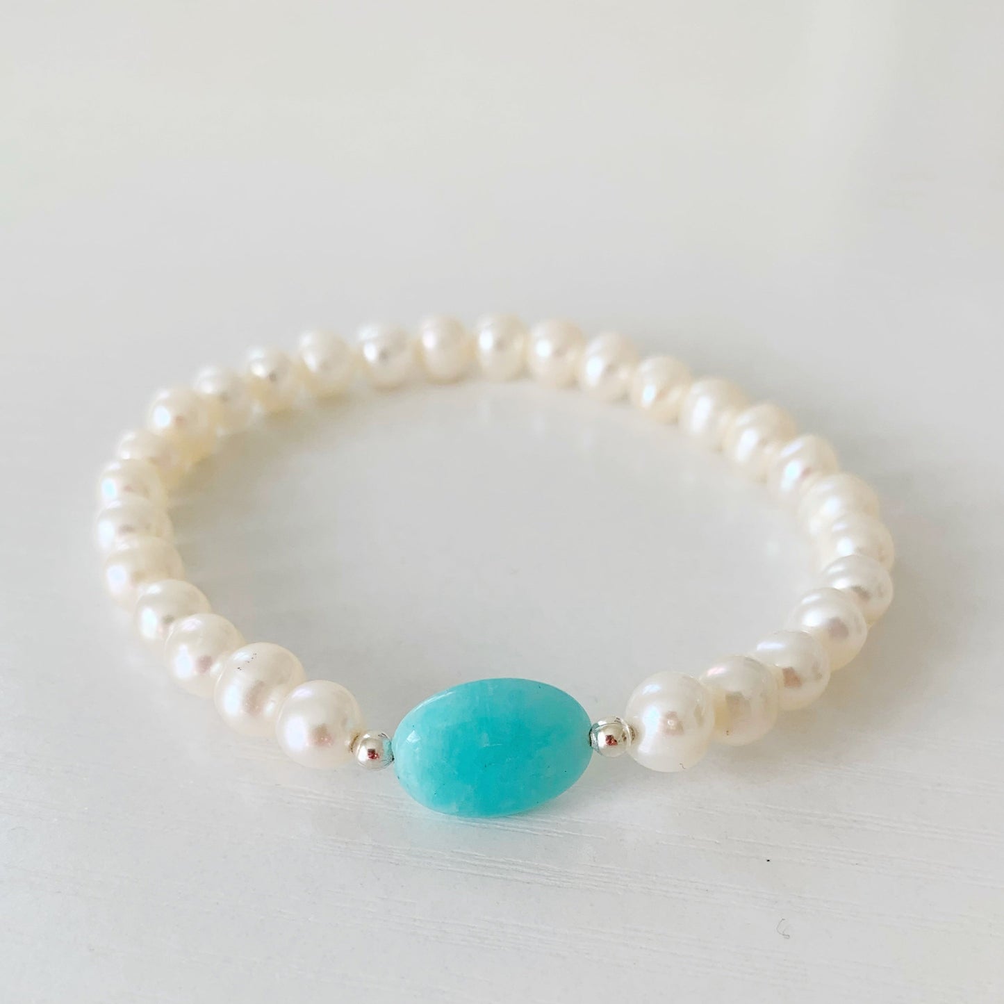 teal sea bracelet by mermaids and madeleines is a stretchy bracelet made with an amazonite oval at the center with sterling silver and freshwater pearls on the rest of it. this bracelet is photographed flat on a white surface