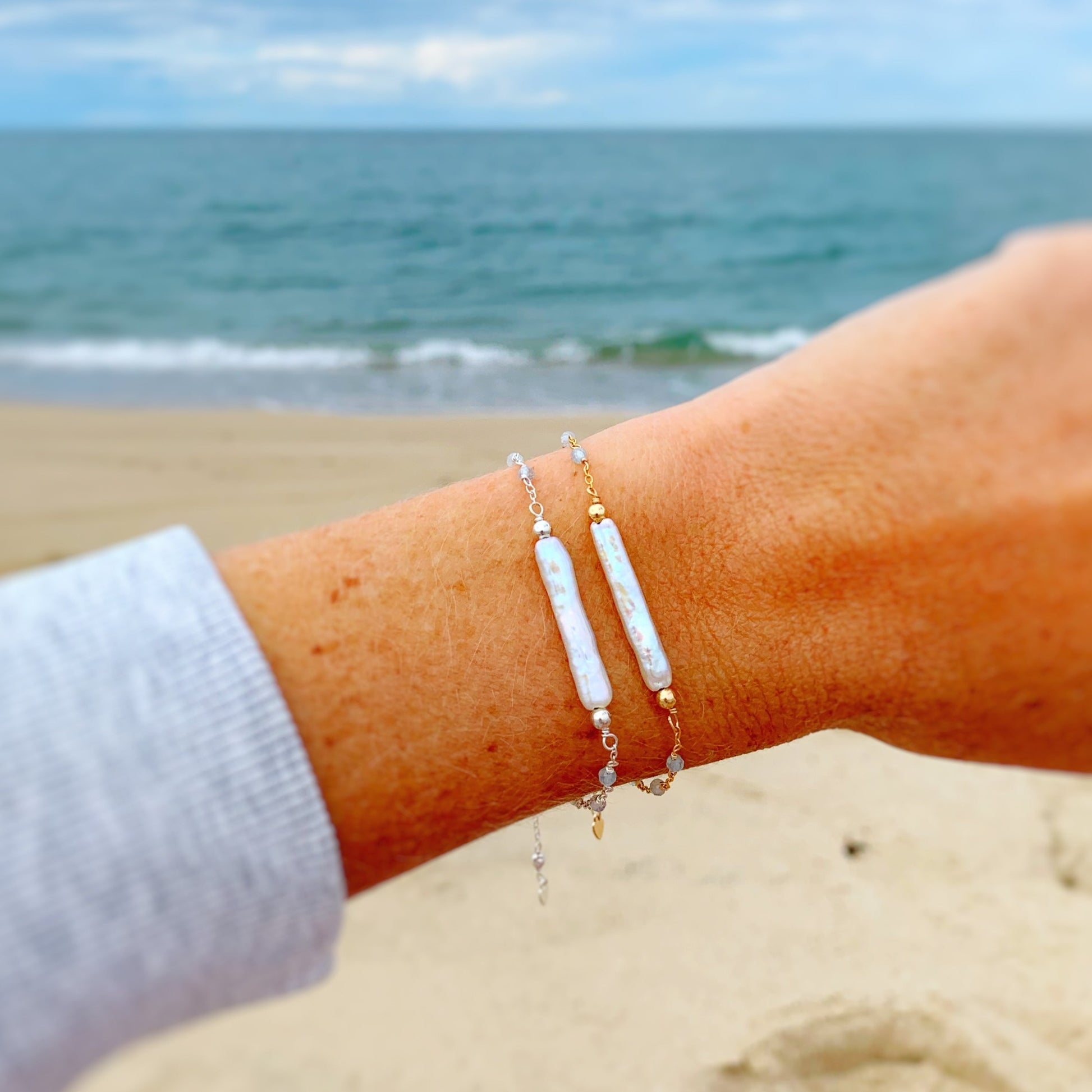 image of a wrist close up with 2 Truro bracelets on. the first is sterling silver and the second is 14k gold filled. the background is a blurry beach scene