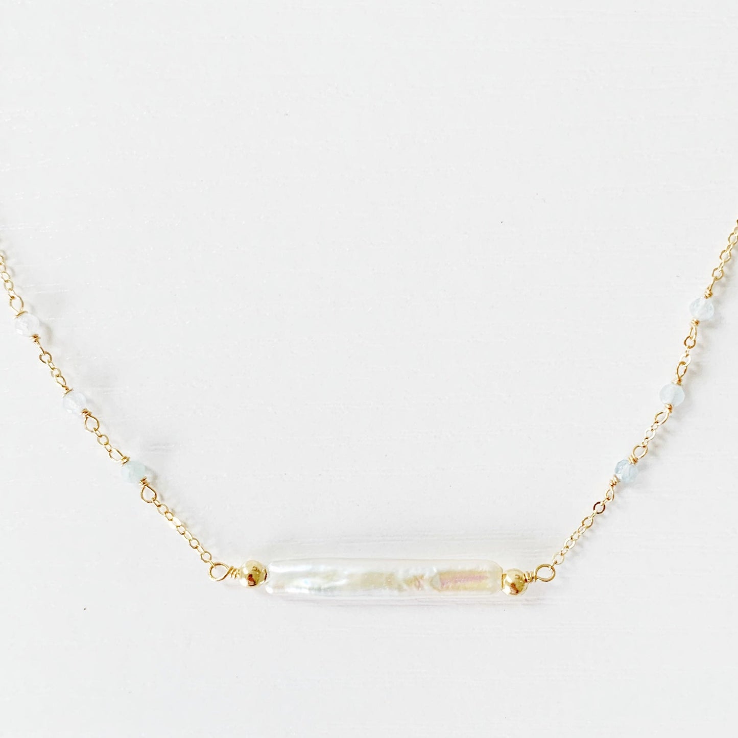 The Truro Necklace by Mermaids and Madeleines has a freshwater stick pearl at the center making a horizontal bar style necklace and has small microfaceted aquamarine beads going up the chain on either side. The chain and findings are 14k gold filled. this necklace has the front middle photographed on a white surface