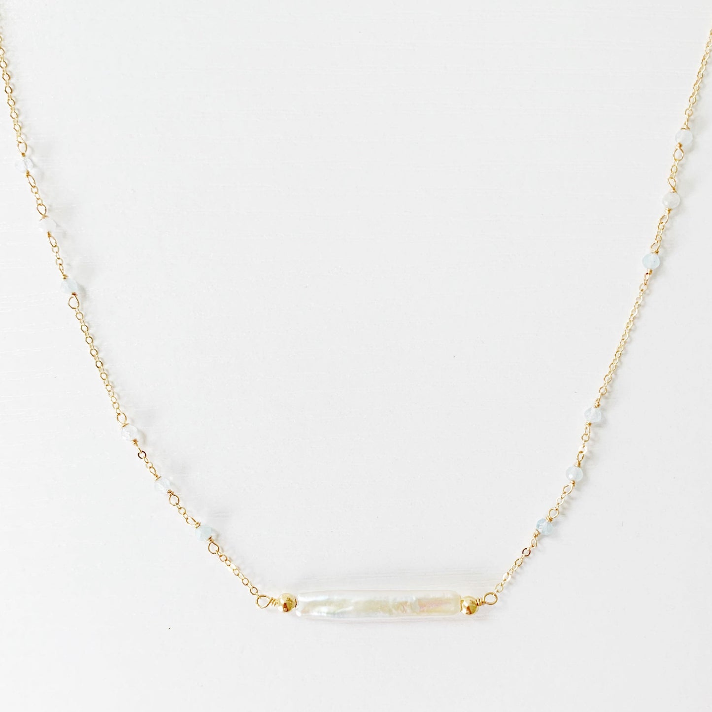 The Truro Necklace by Mermaids and Madeleines has a freshwater stick pearl at the center making a horizontal bar style necklace and has small microfaceted aquamarine beads going up the chain on either side. The chain and findings are 14k gold filled. this necklace has the front half photographed on a white surface