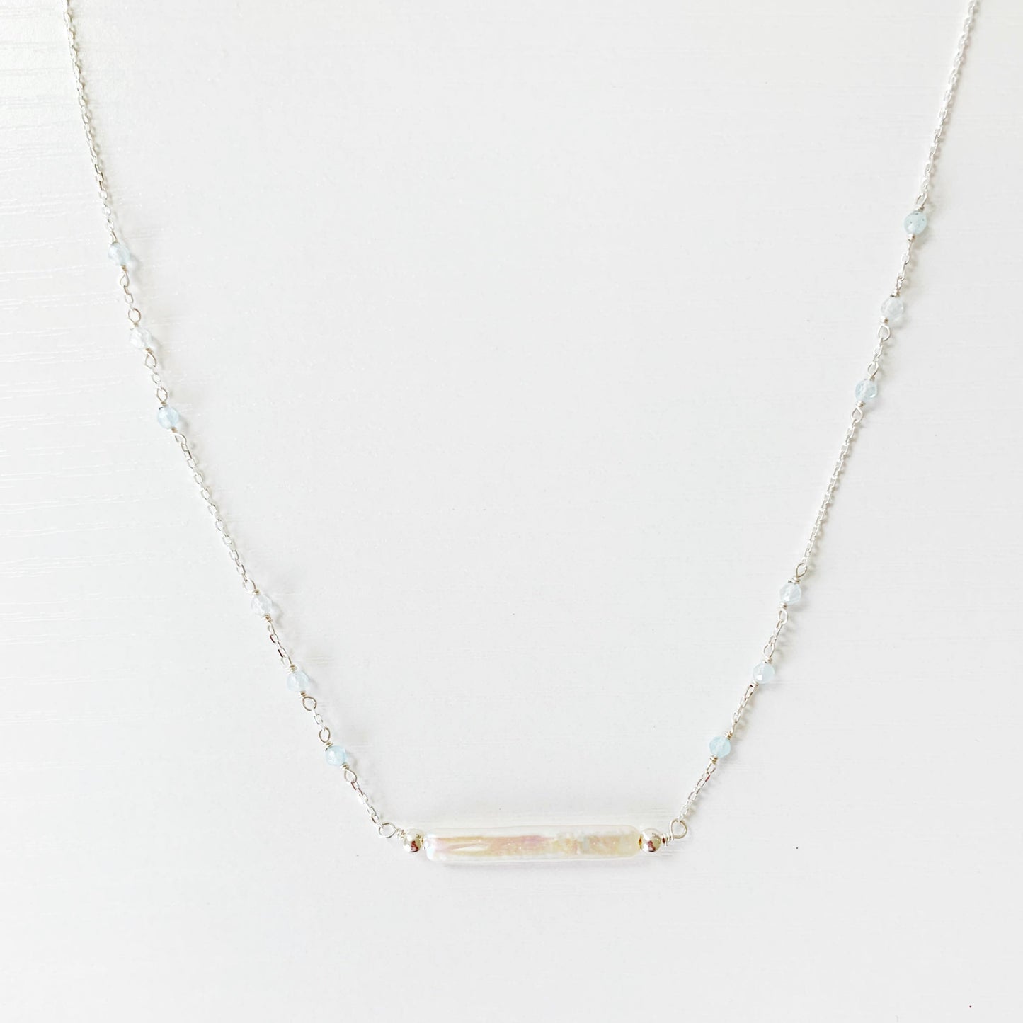 The Truro Necklace by Mermaids and Madeleines features a freshwater stick pearl at the center like a horizontal bar necklace with tiny microfaceted aquamarine beads going up the chain on either side. the chain and findings are sterling silver. this necklace is photographed on a white surface