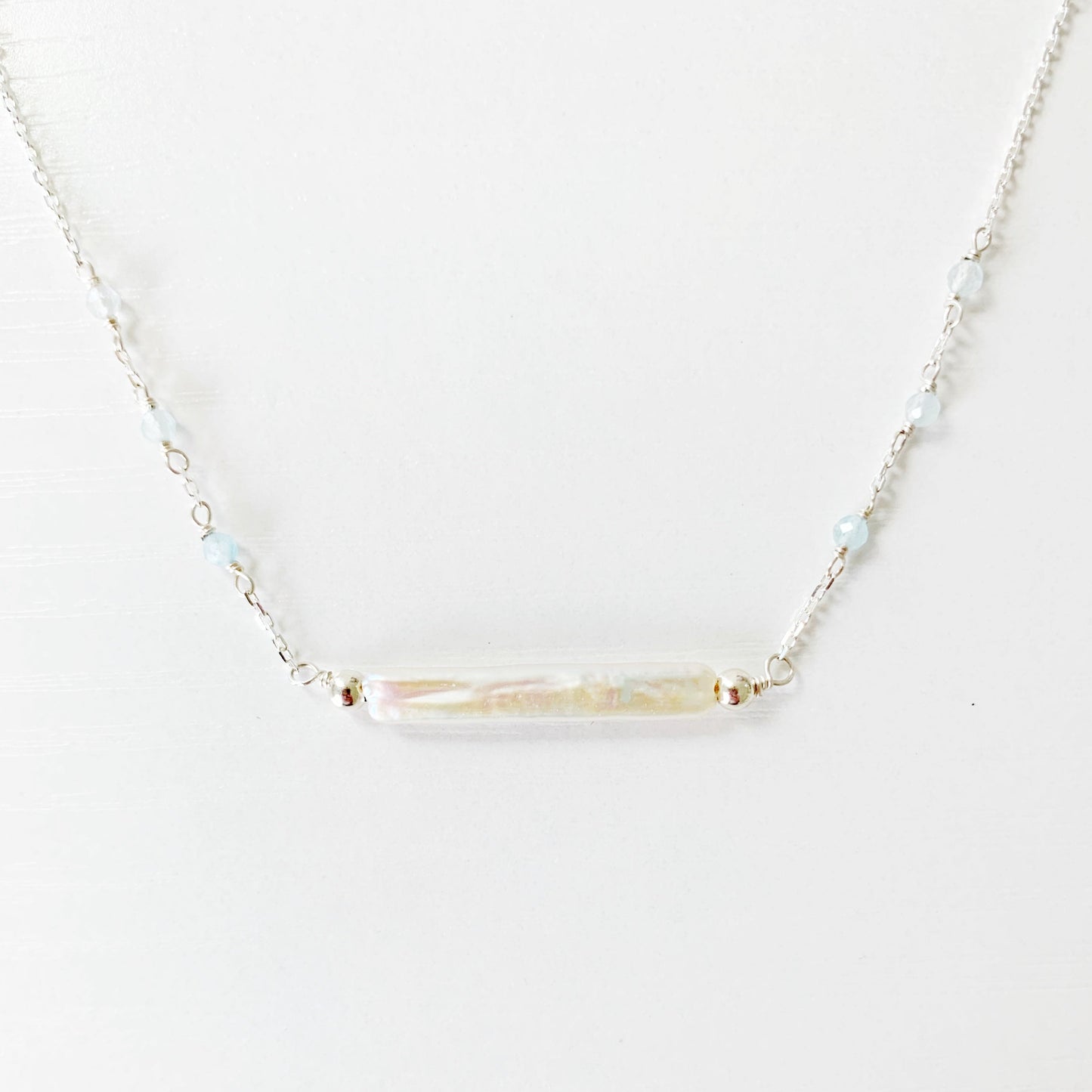 The Truro Necklace by Mermaids and Madeleines features a freshwater stick pearl at the center like a horizontal bar necklace with tiny microfaceted aquamarine beads going up the chain on either side. the chain and findings are sterling silver. this necklace is photographed with just the very front center of the necklace showing and its on a white surface