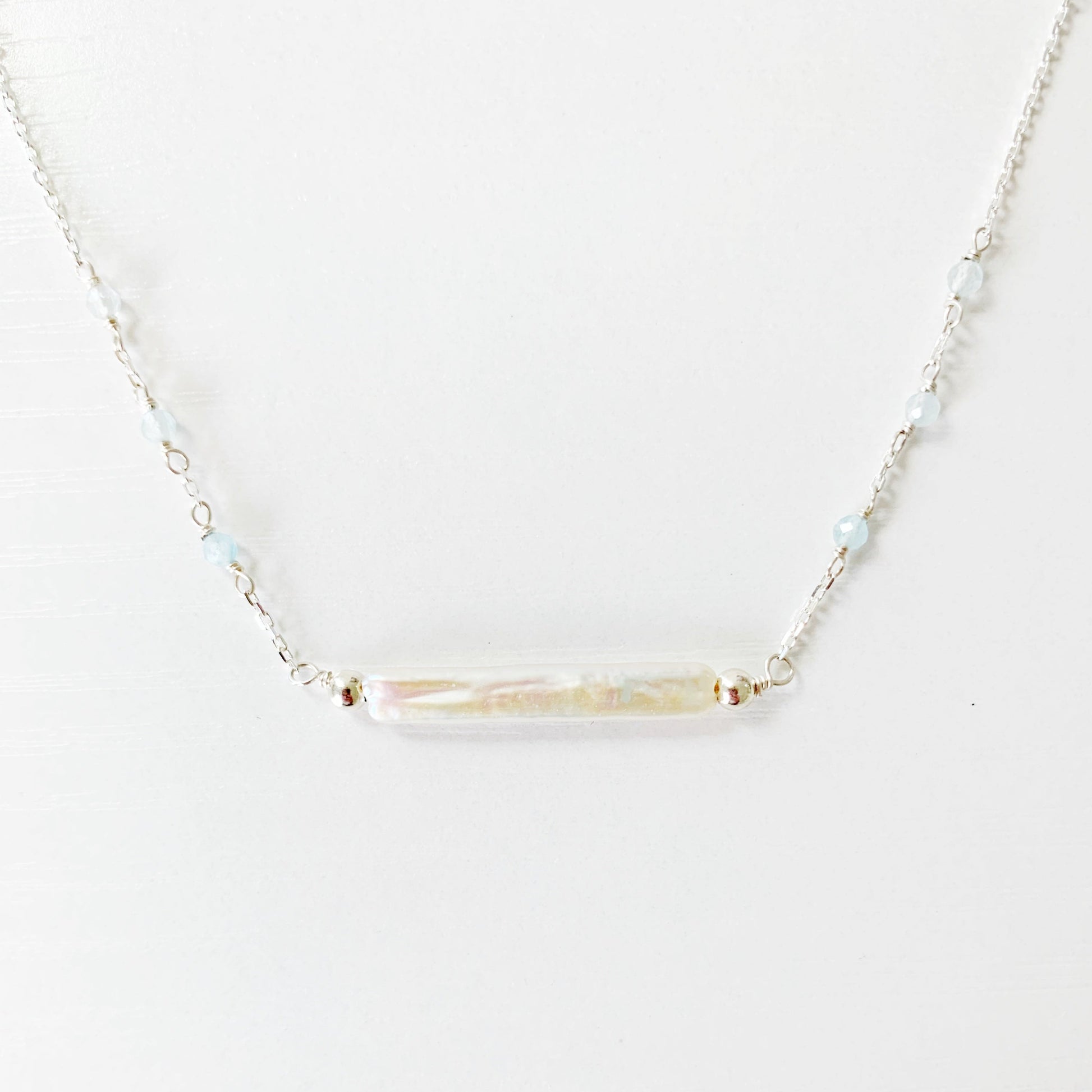 The Truro Necklace by Mermaids and Madeleines features a freshwater stick pearl at the center like a horizontal bar necklace with tiny microfaceted aquamarine beads going up the chain on either side. the chain and findings are sterling silver. this necklace is photographed with just the very front center of the necklace showing and its on a white surface