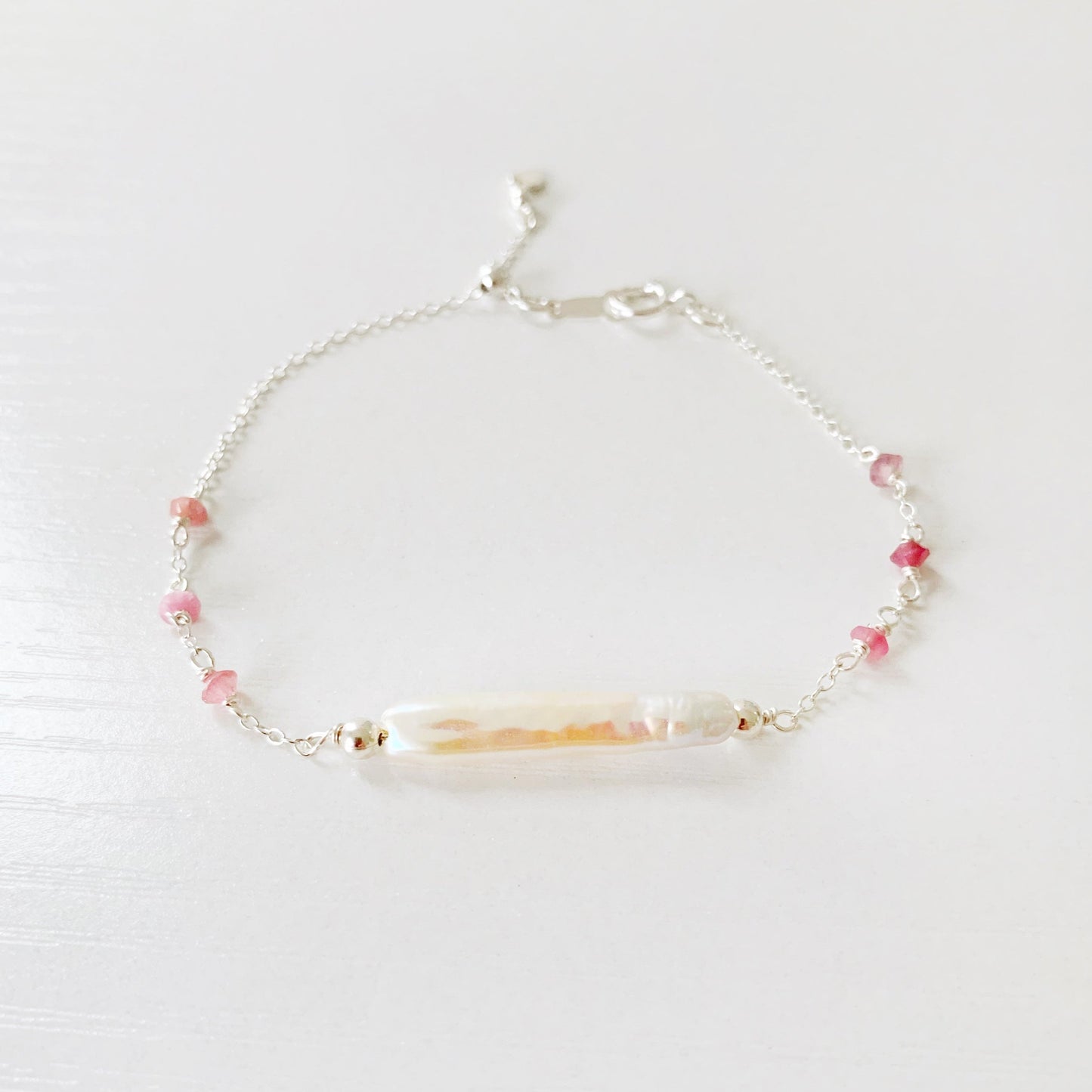 the pink azalea adjustable bracelet by mermaids and madeleines is a sterling silver chain bracelet with a slide bead near the closure. the center of the bracelet features a freshwater pearl bar and it has small pink tourmaline beads on the sides of the chain. this bracelet is photographed on a white surface with a front view