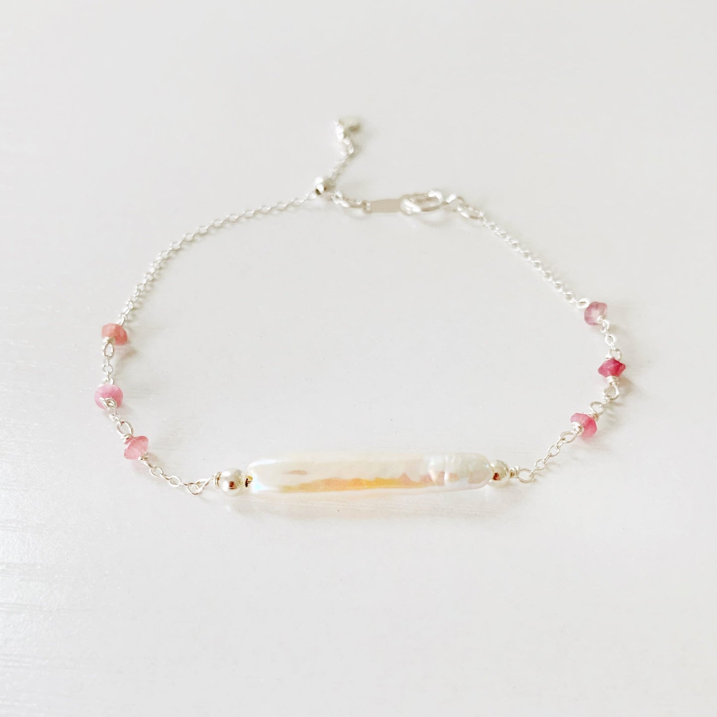 the pink azalea adjustable bracelet by mermaids and madeleines is a sterling silver chain bracelet with a slide bead near the closure. the center of the bracelet features a freshwater pearl bar and it has small pink tourmaline beads on the sides of the chain. this bracelet is photographed on a white surface with a front view