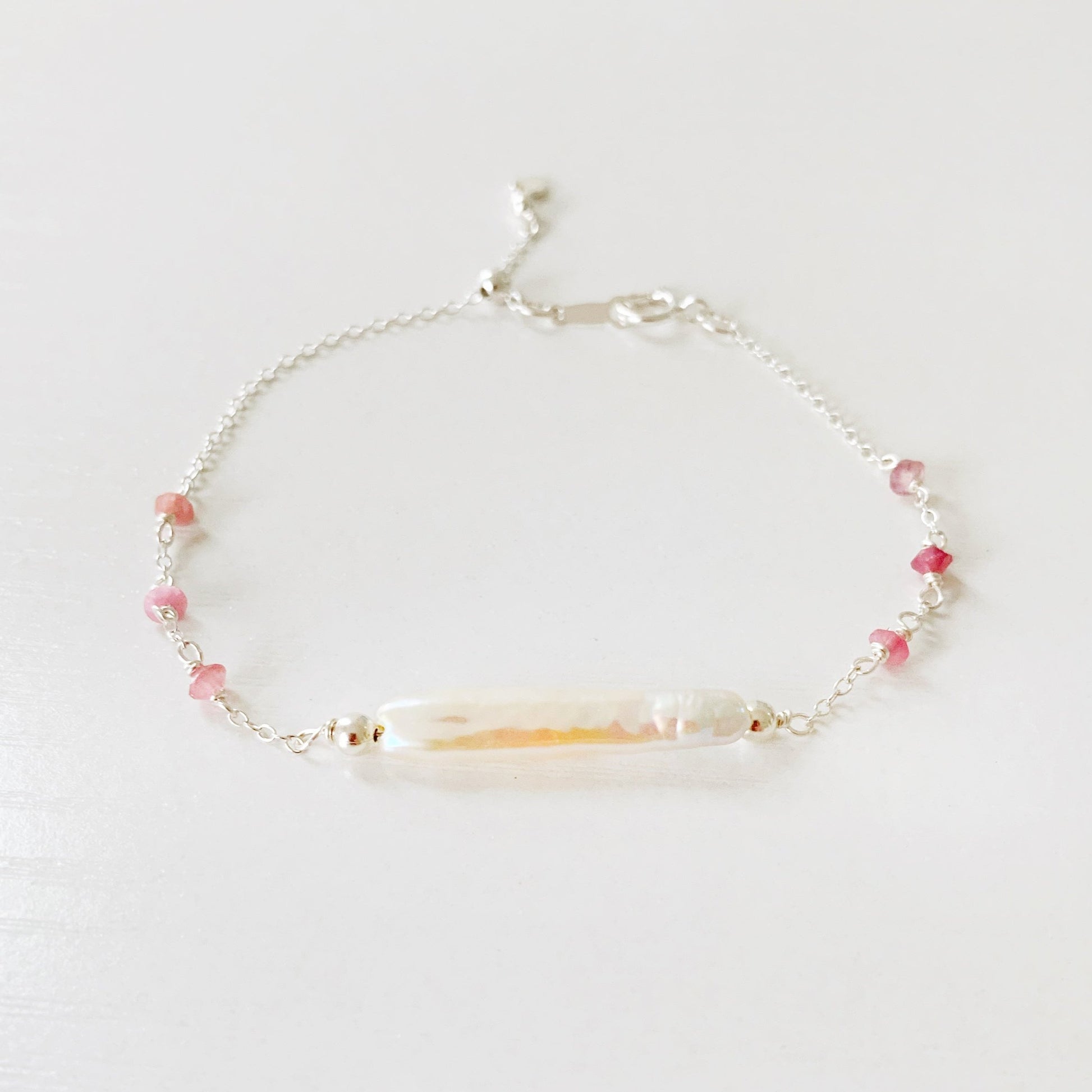 the pink azalea adjustable bracelet by mermaids and madeleines is a sterling silver chain bracelet with a slide bead near the closure. the center of the bracelet features a freshwater pearl bar and it has small pink tourmaline beads on the sides of the chain. this bracelet is photographed on a white surface with a front view