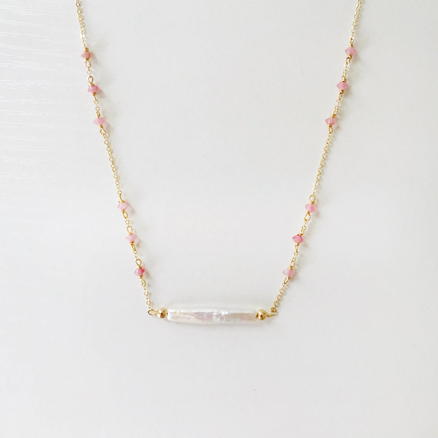 the pink azalea necklace by mermaids and madeleines is a freshwater pearl bar necklace with pink tourmaline faceted beads in groups of 3 on the 14k gold filled chain. this necklace is photographed with the front half in view on a white surface.