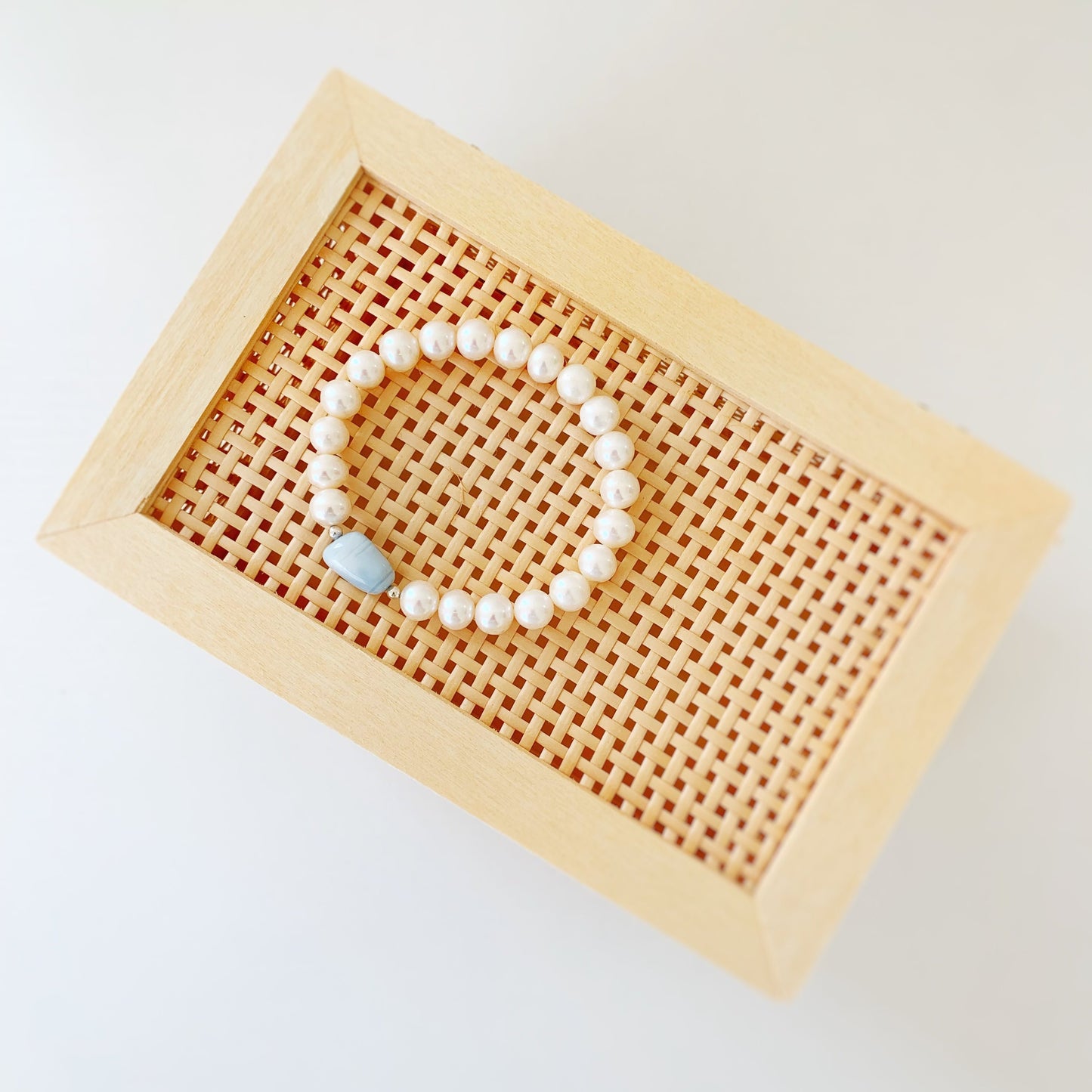 the blue hydrangea bracelet by mermaids and madeleines is a stretch bracelet with a pebble shaped blue opal in the center with freshwater pearls on the rest of the bracelet. this one is photographed in a flatlay on top of a woven box on a white table