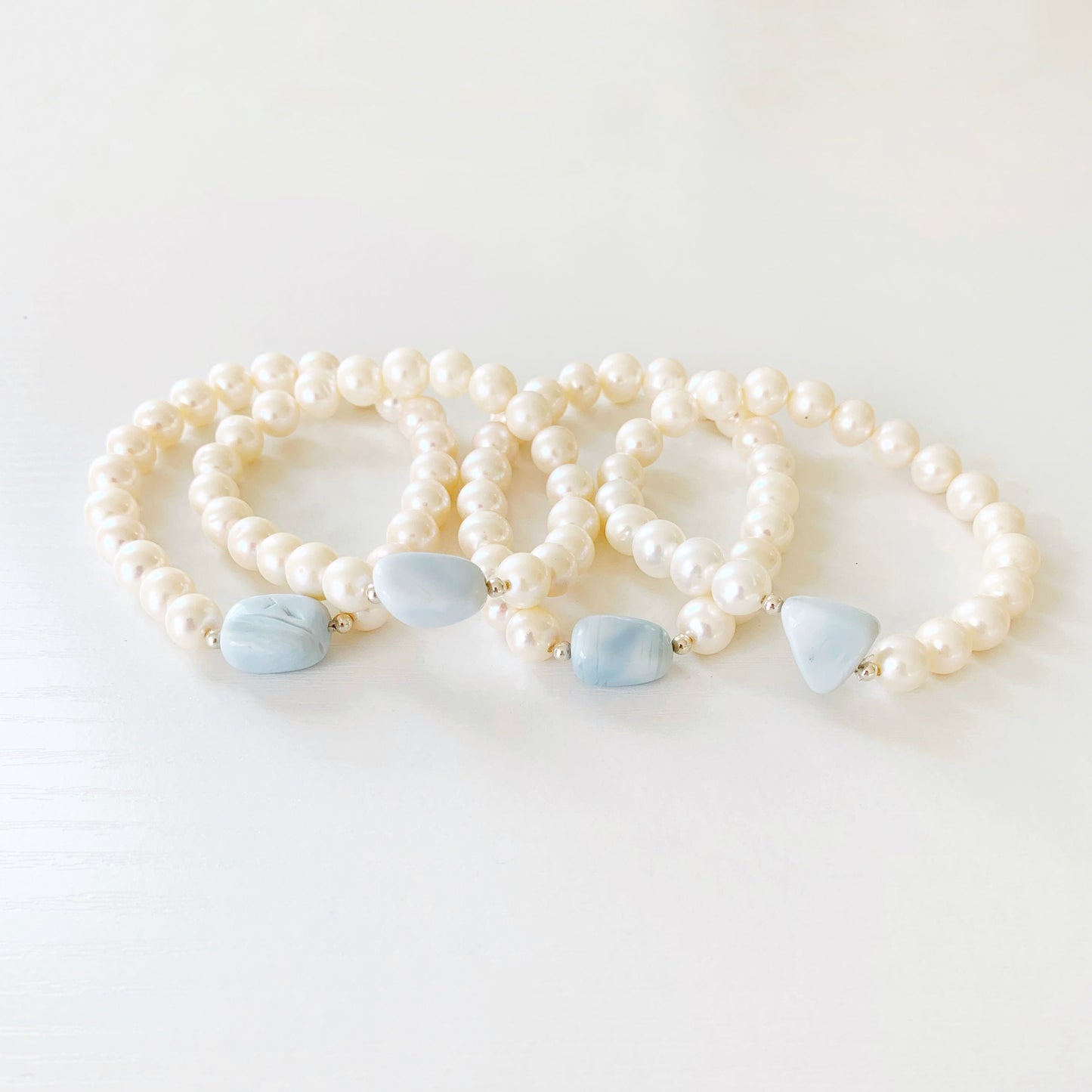 the blue hydrangea bracelet by mermaids and madeleines is a freshwater pearl stretch bracelet with a blue opal pebble shaped bead at the center. this is a photograph of 4 of the blue hydrangea bracelets grouped on a white table