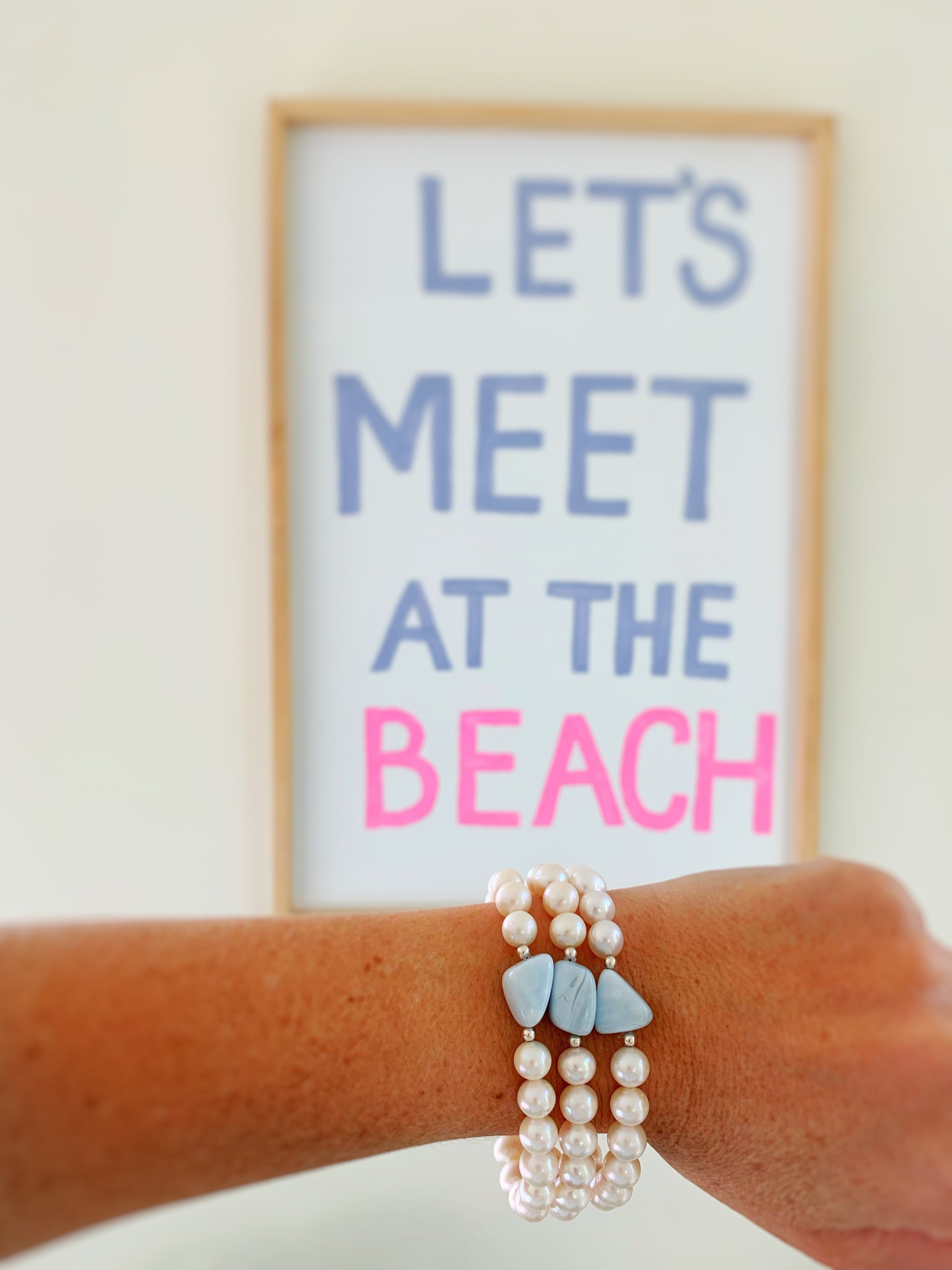 the blue hydrangea bracelet by mermaids and madeleines is a freshwater pearl stretch bracelet with a blue opal pebble bead at the center. this is a photograph of 3 blue hydrangea bracelets on a wrist with a beach text poster in the background.