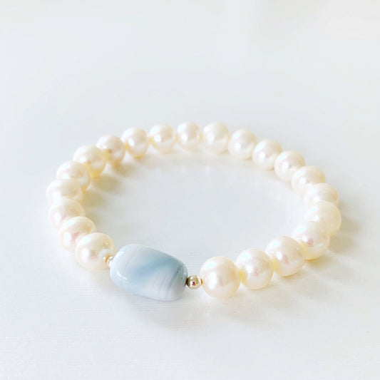 the blue hydrangea stretch bracelet by mermaids and madeleines is designed with a blue opal pebble bead at the center with freshwater pearls along the rest of the bracelet. this bracelet is photographed flat on a white surface