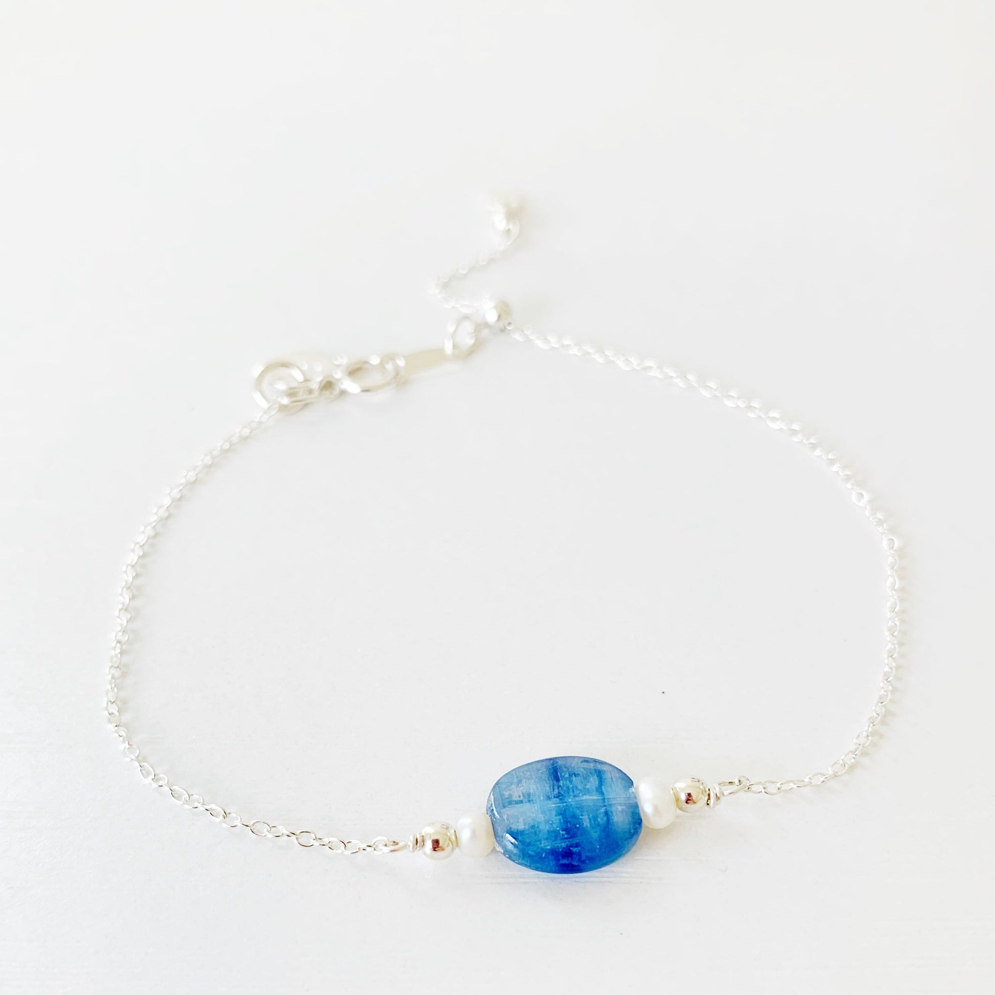 The brewster bracelet by mermaids and madeleines is an adjustable chain bracelet with an oval blue kyanite bead at the center with 2 small pearls on each side. there is a slide bead near the clasp and the chain and findings are sterling silver. this bracelet is photographed on a white surface