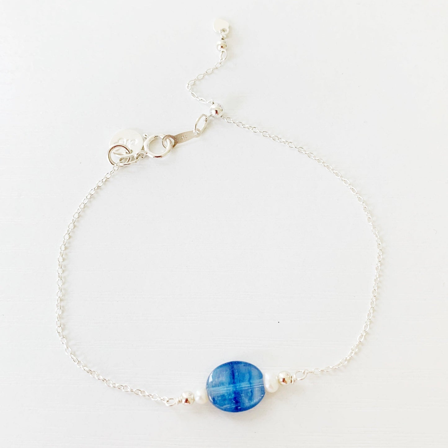 The brewster bracelet by mermaids and madeleines is an adjustable chain bracelet with an oval blue kyanite bead at the center with 2 small pearls on each side. there is a slide bead near the clasp and the chain and findings are 14k gold filled. this bracelet is photographed on a white surface