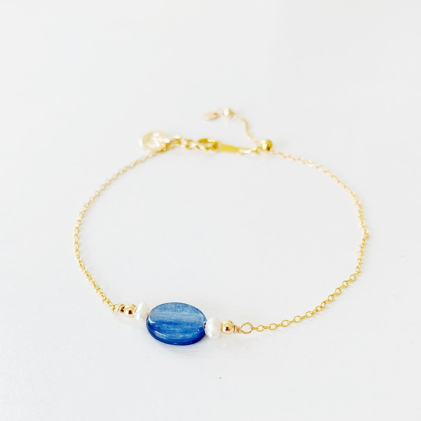 The brewster bracelet by mermaids and madeleines is an adjustable chain bracelet with an oval blue kyanite bead at the center with 2 small pearls on each side. there is a slide bead near the clasp and the chain and findings are 14k gold filled. this bracelet is photographed on a white surface