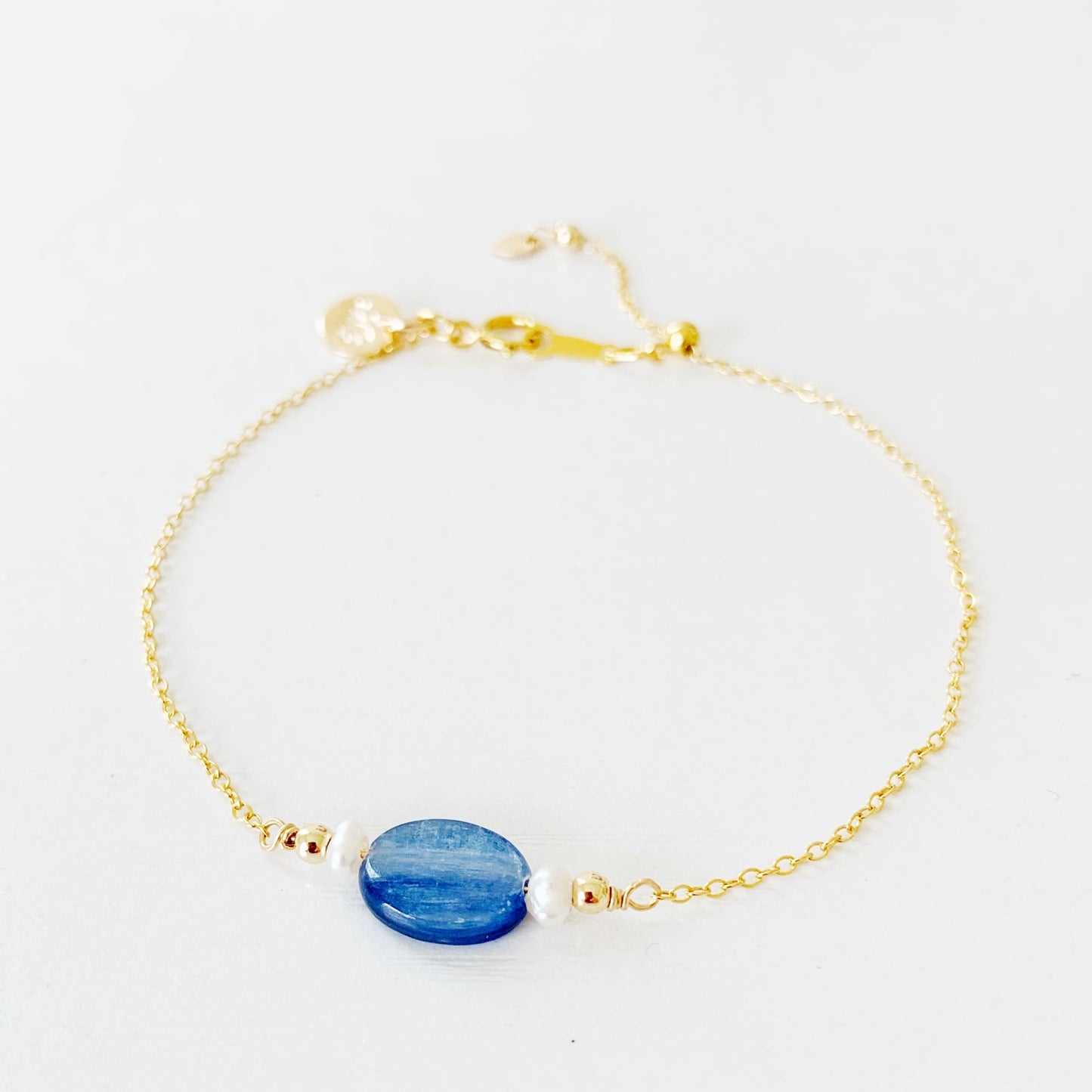 The brewster bracelet by mermaids and madeleines is an adjustable chain bracelet with an oval blue kyanite bead at the center with 2 small pearls on each side. there is a slide bead near the clasp and the chain and findings are 14k gold filled. this bracelet is photographed on a white surface