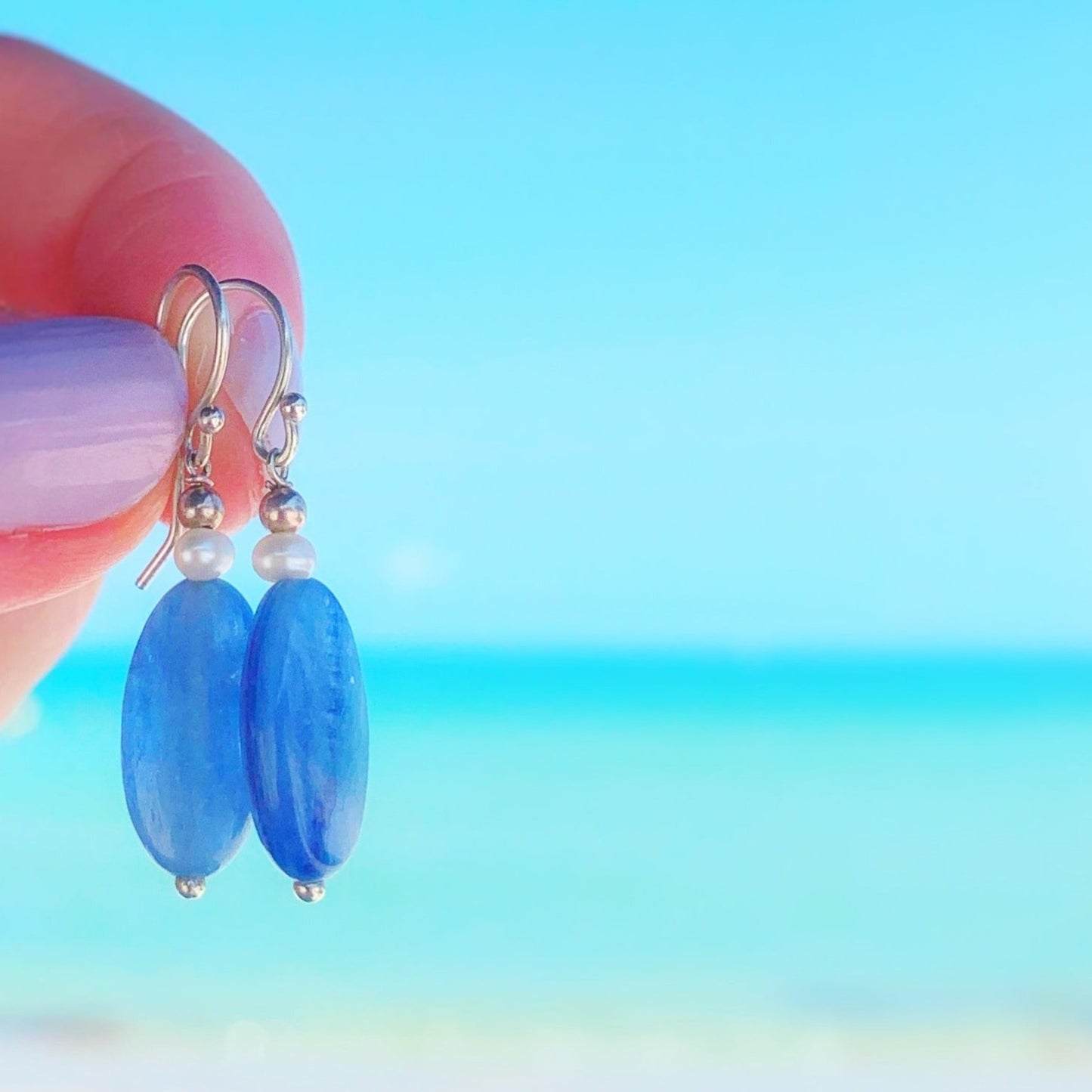 Brewster earrings by mermaids and madeleines are oblong blue kyanite bead drop earrings with petite freshwater pearls on top and sterling silver findings. this pair is held up in a hand with a beach background