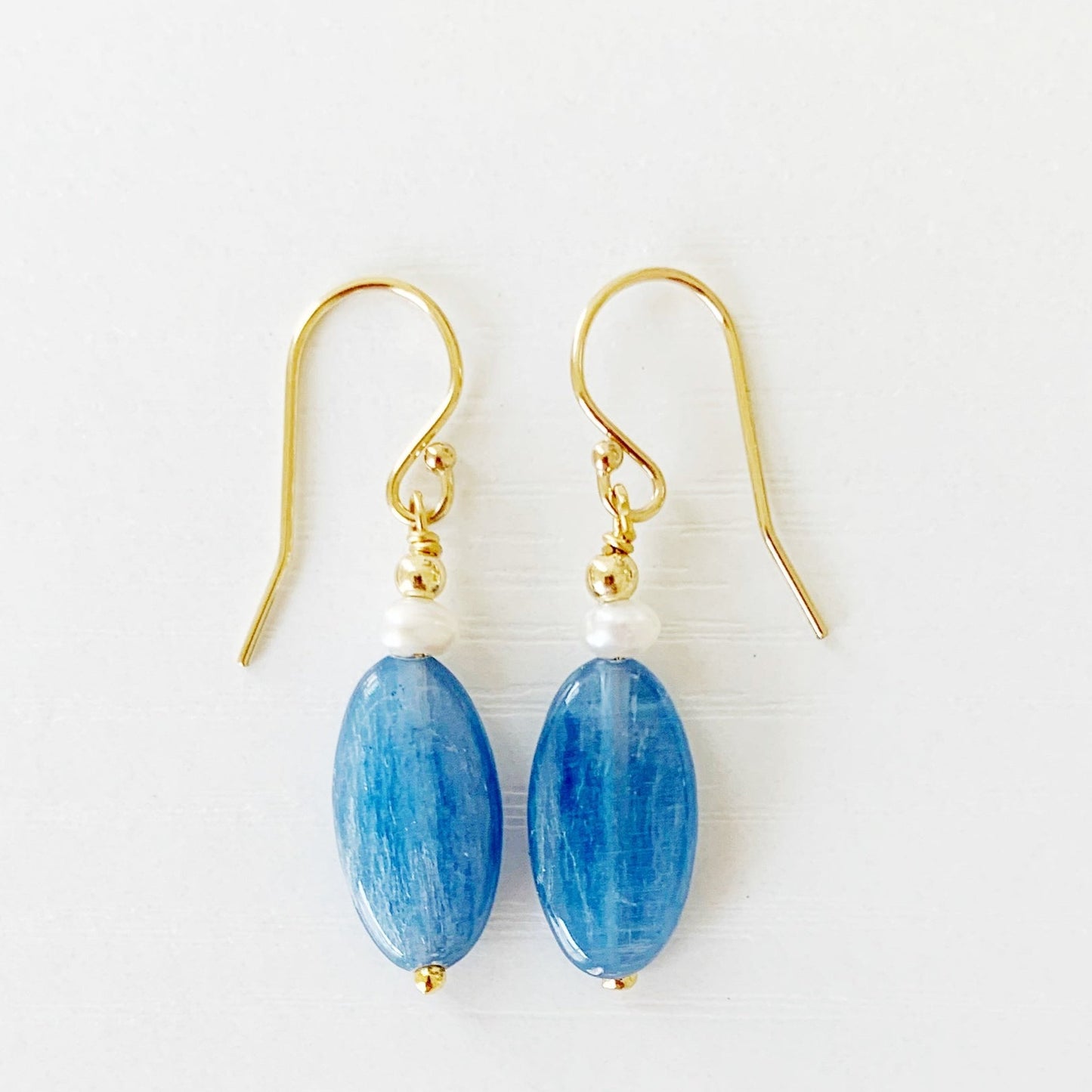 the brewster earrings by mermaids and madeleines are long oval blue kyanite beads with small pearls on top before the 14k gold filled findings. this pair is photographed flat on a white surface