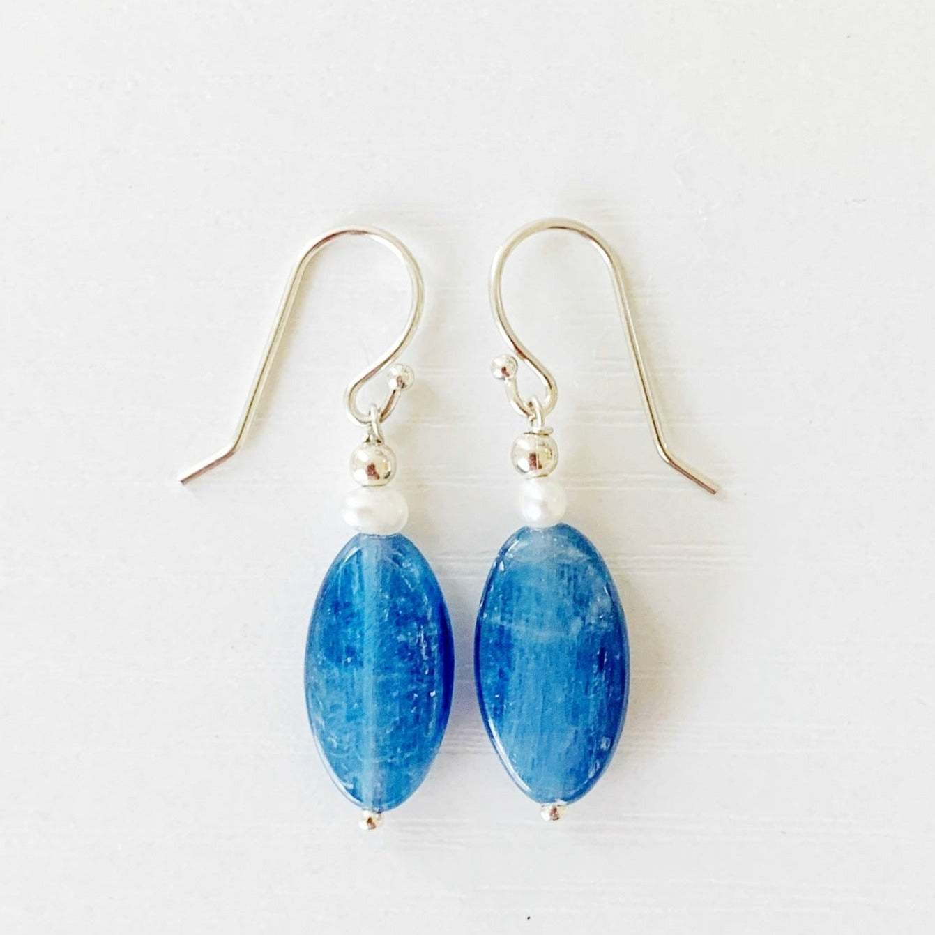 The brewster earrings by mermaids and madeleines are long oval blue kyanite drops with a small pearl near the ear wire. the findings are sterling silver. this pair of earrings is photographed flat on a white surface