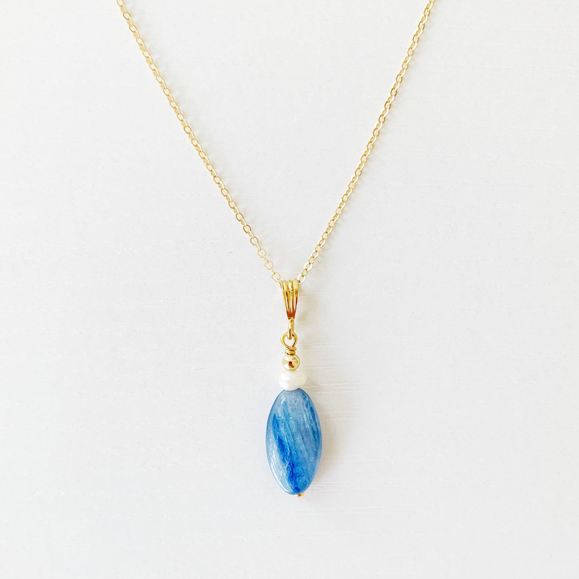 the brewster necklace by mermaids and madeleines is a pendant style necklace with a long oval blue kyanite bead dropped from a 14k gold filled chain and findings. this necklace is photographed flat on a white surface